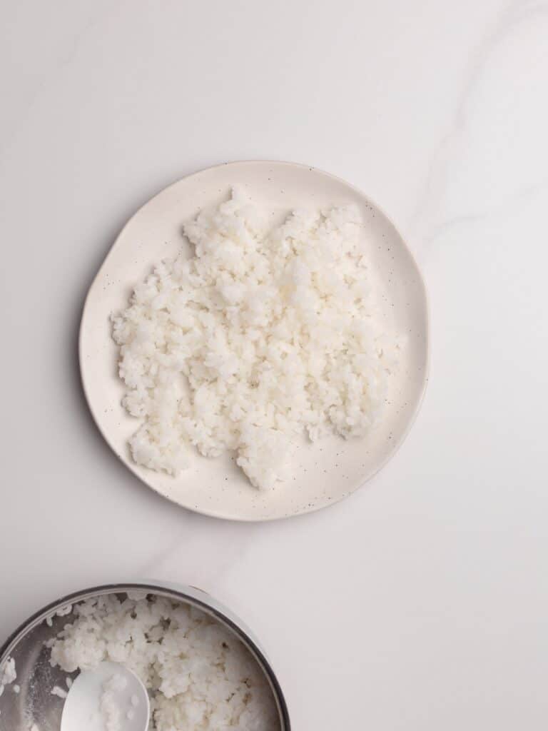 A white plate with a serving of cooked white rice sits on a white surface. Part of a metal pot with more rice and a spoon is visible in the bottom left corner.