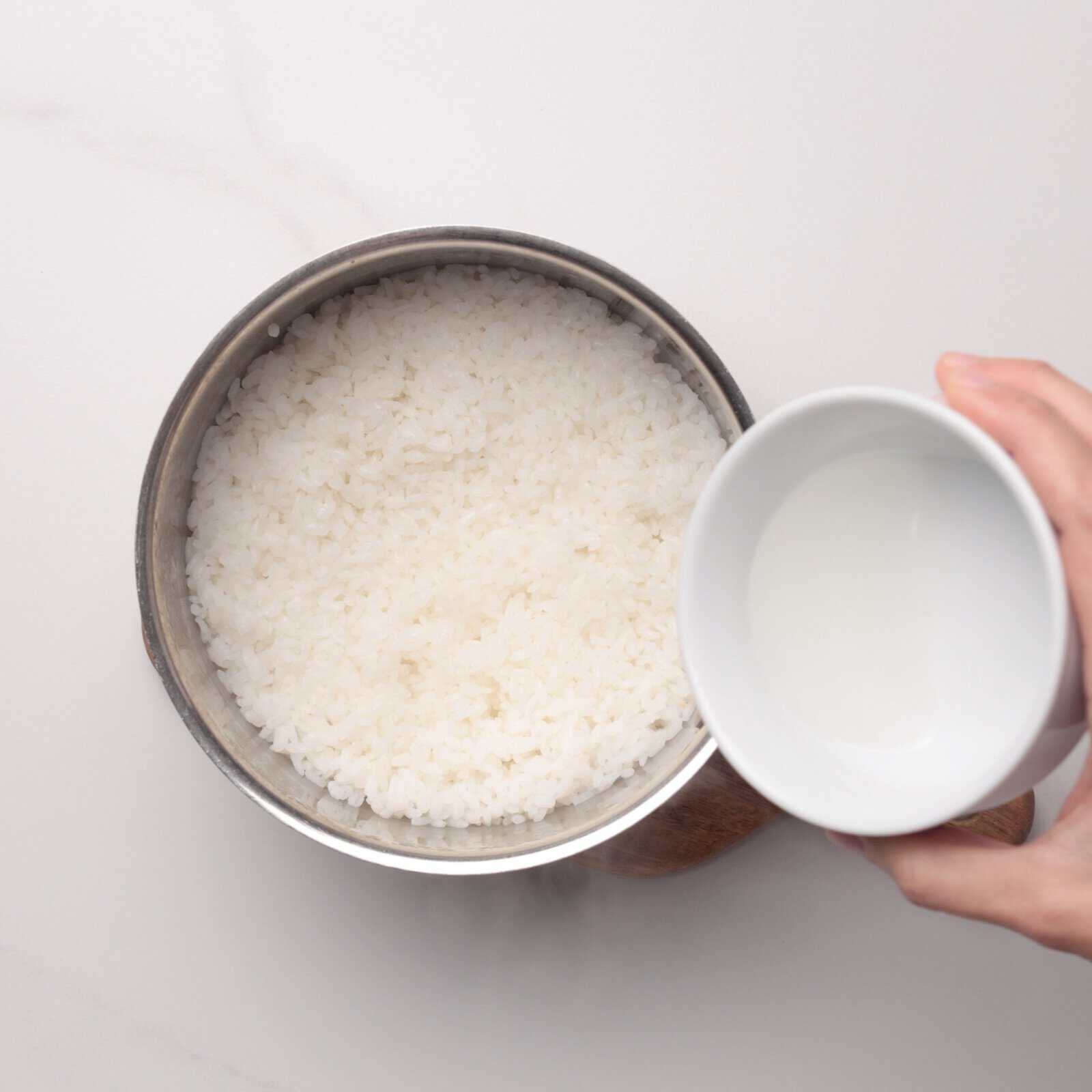 A hand holds a white cup of water above a pot of cooked white rice on a white surface, preparing to pour the water into the rice.