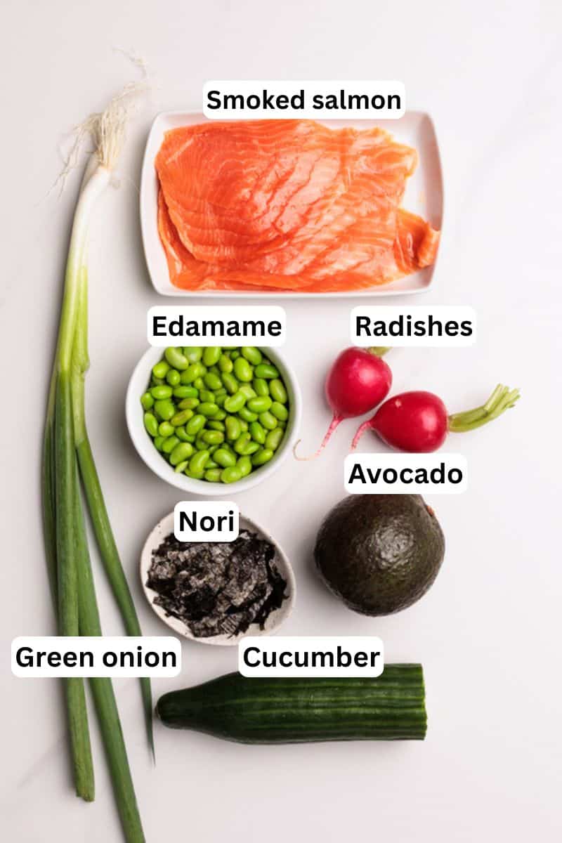 A flat lay of sushi bowl ingredients: smoked salmon on a plate, edamame in a bowl, two radishes, an avocado, a bowl of nori, green onion stalks, and a cucumber, all labeled.