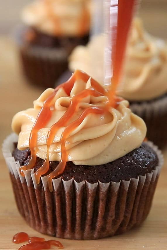 A chocolate cupcake with swirled caramel frosting is being drizzled with caramel sauce, with another similarly decorated cupcake blurred in the background.
