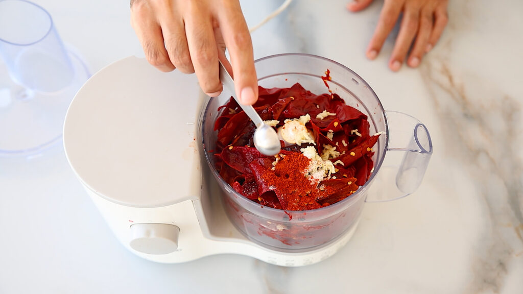 A hand uses a small spoon to add spices to a food processor containing red chili peppers, garlic, and other ingredients on a white countertop.