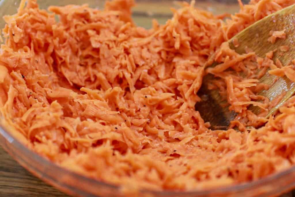 A close-up of a glass bowl filled with shredded sweet potato being mixed with a wooden spoon.