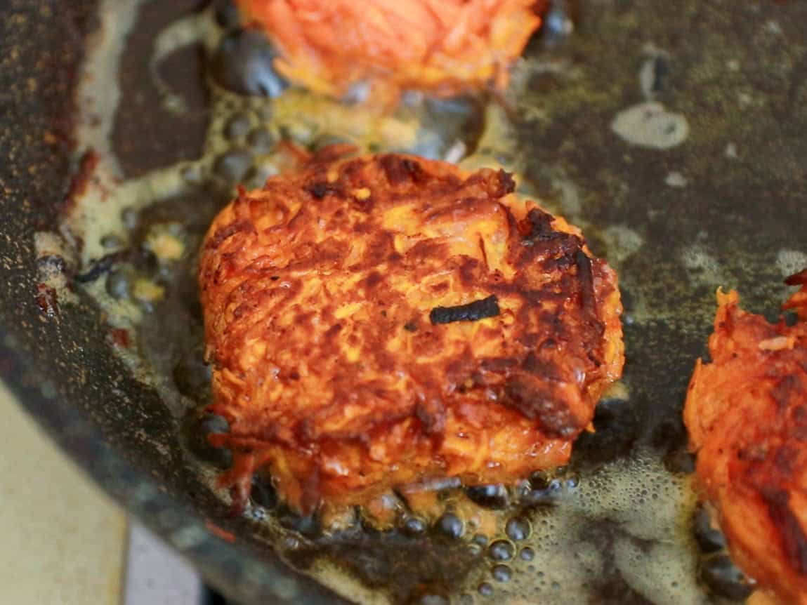 A sweet potato patty is frying in a pan with bubbling oil, showing a crispy, browned surface and orange interior. Other patties are partially visible nearby.