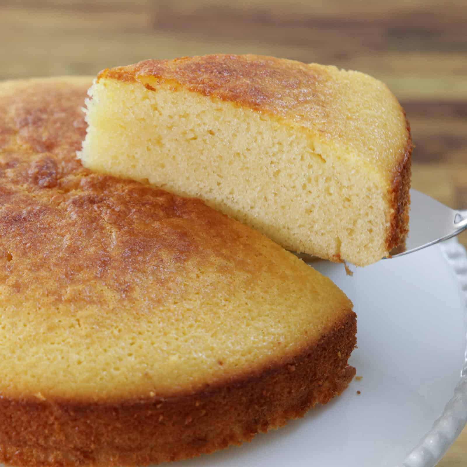 A close-up of a golden, moist homemade 4-ingredient condensed milk cake with a slice being lifted from the whole cake on a white plate. The cake has a slightly browned edge and a soft, fluffy texture inside.