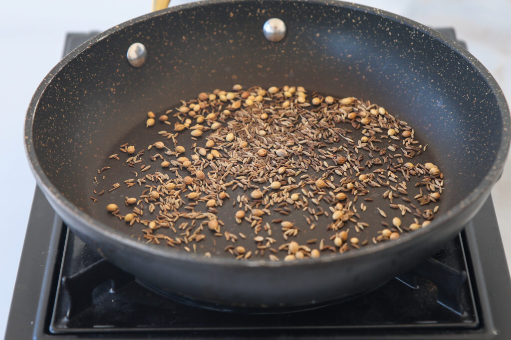 A close-up of a black frying pan on a stove, containing whole coriander and cumin seeds being dry roasted. The seeds are scattered and lightly toasted.