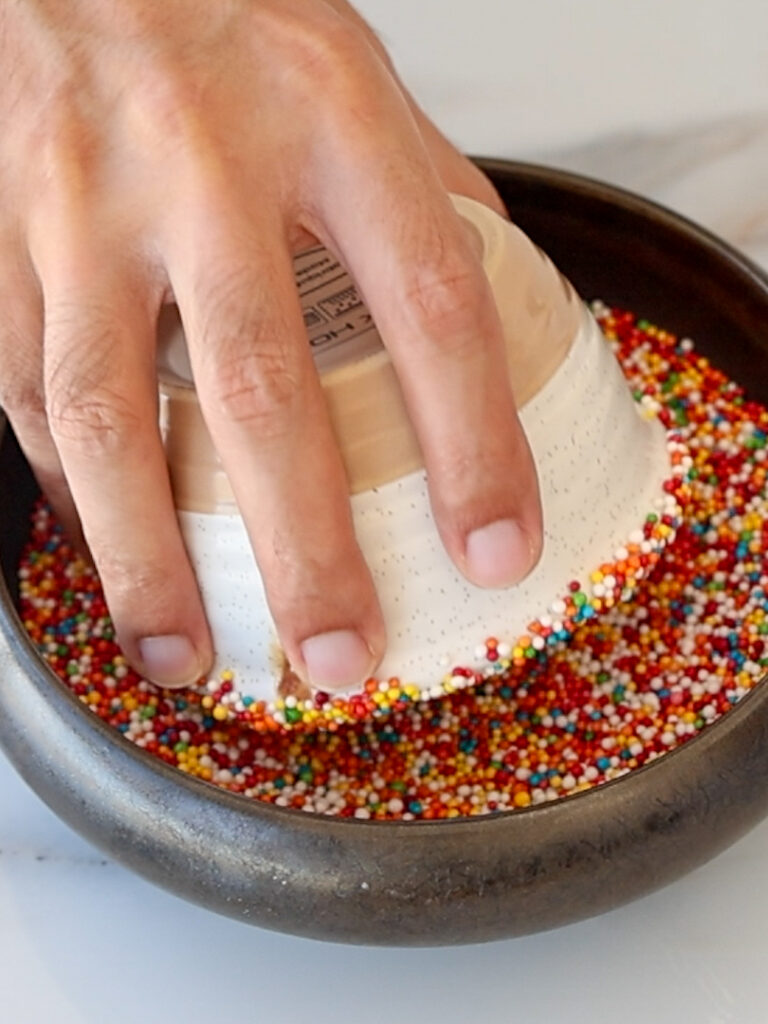 A hand presses an upside-down cup with a coated rim into a bowl filled with colorful round sprinkles, decorating the edge of the cup.
