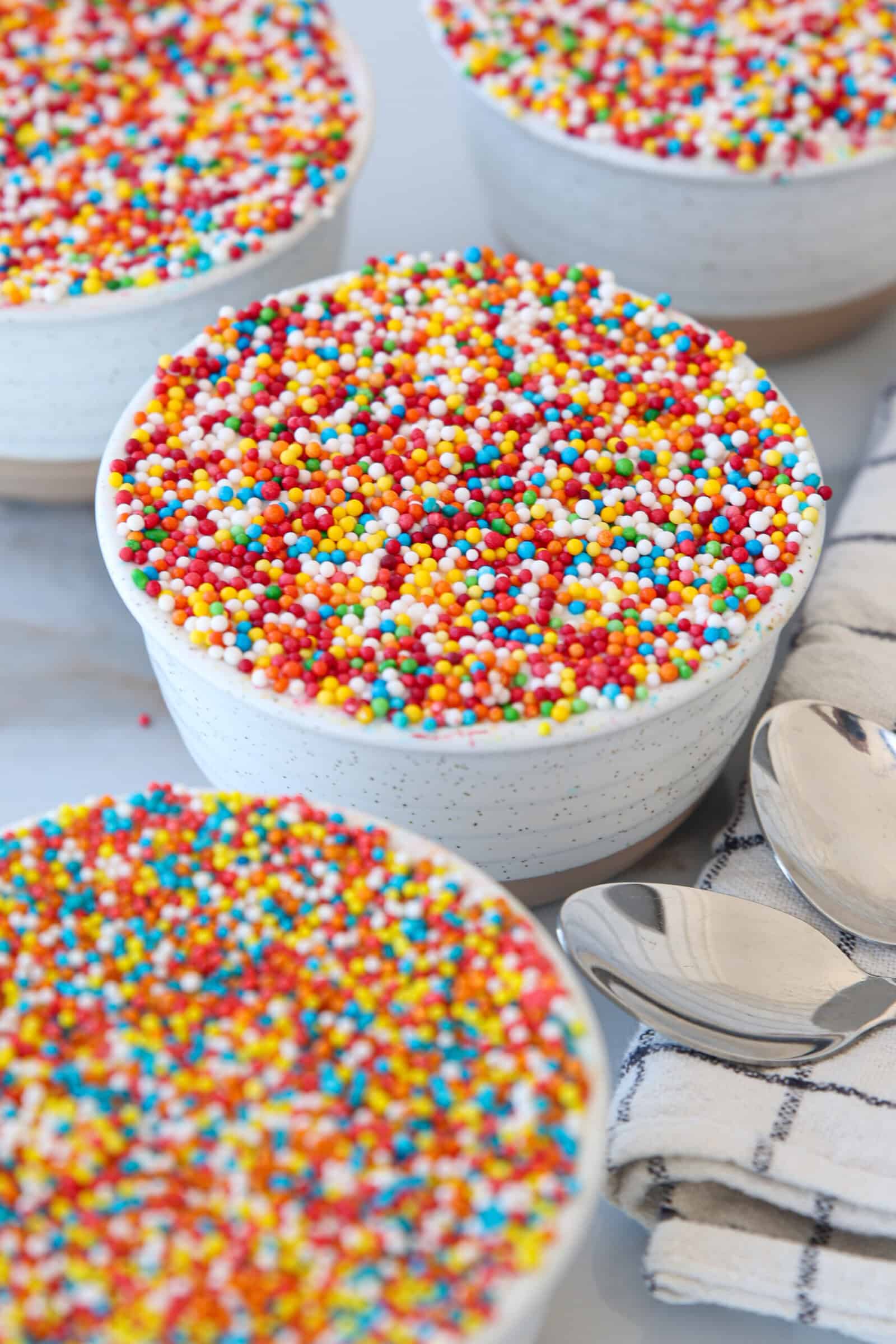Four white ramekins filled with the viral dot cakes and topped with colorful round sprinkles, set on a white surface next to a striped cloth napkin and two silver spoons.