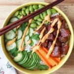 A bowl of rice topped with sliced cucumber, edamame, carrot sticks, marinated fish, parsley, and orange sauce, with chopsticks resting on the bowl.