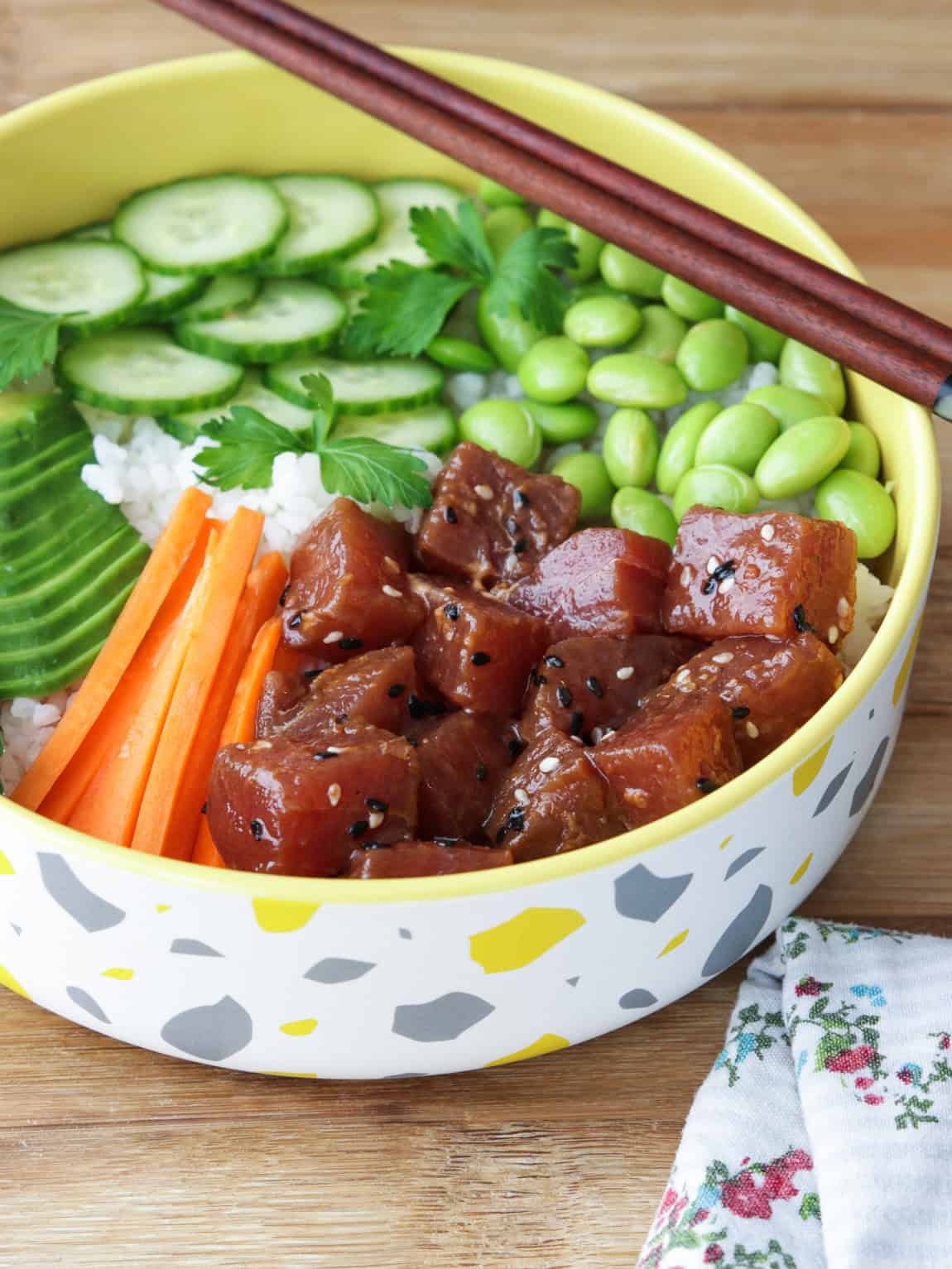 A colorful poke bowl with tuna, carrots, avocado, cucumber, edamame, rice, parsley, and chopsticks on top, in a decorative bowl on a wooden table next to a floral napkin.