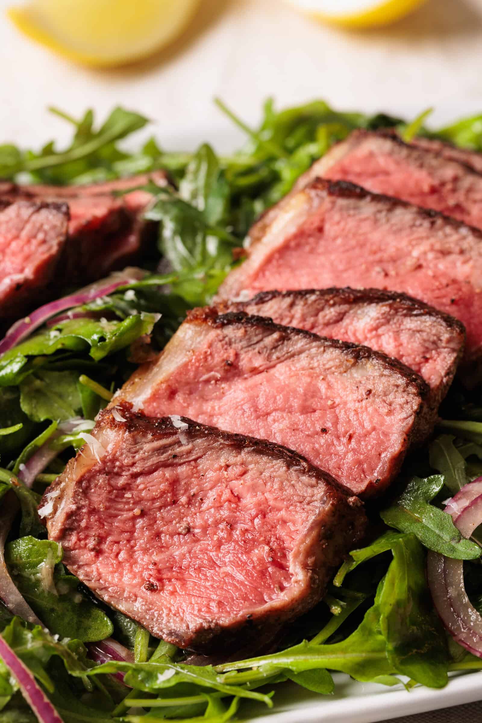 Five slices of medium-rare steak are arranged on a bed of fresh arugula salad, with a blurred lemon wedge in the background. The steak has a seared crust and a juicy pink center.