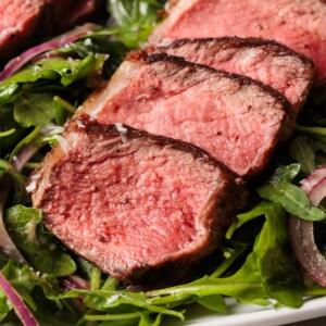 Close-up of three slices of medium-rare steak with a seared crust, served on a bed of fresh arugula and red onion salad.