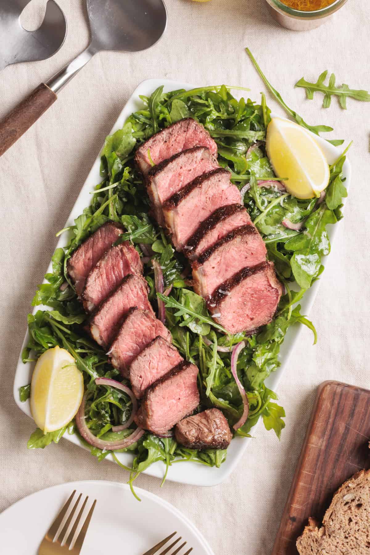 Arugula Steak Salad with Sliced steak served on a bed of arugula and red onions, garnished with lemon wedges, presented on a white rectangular plate with serving utensils nearby.