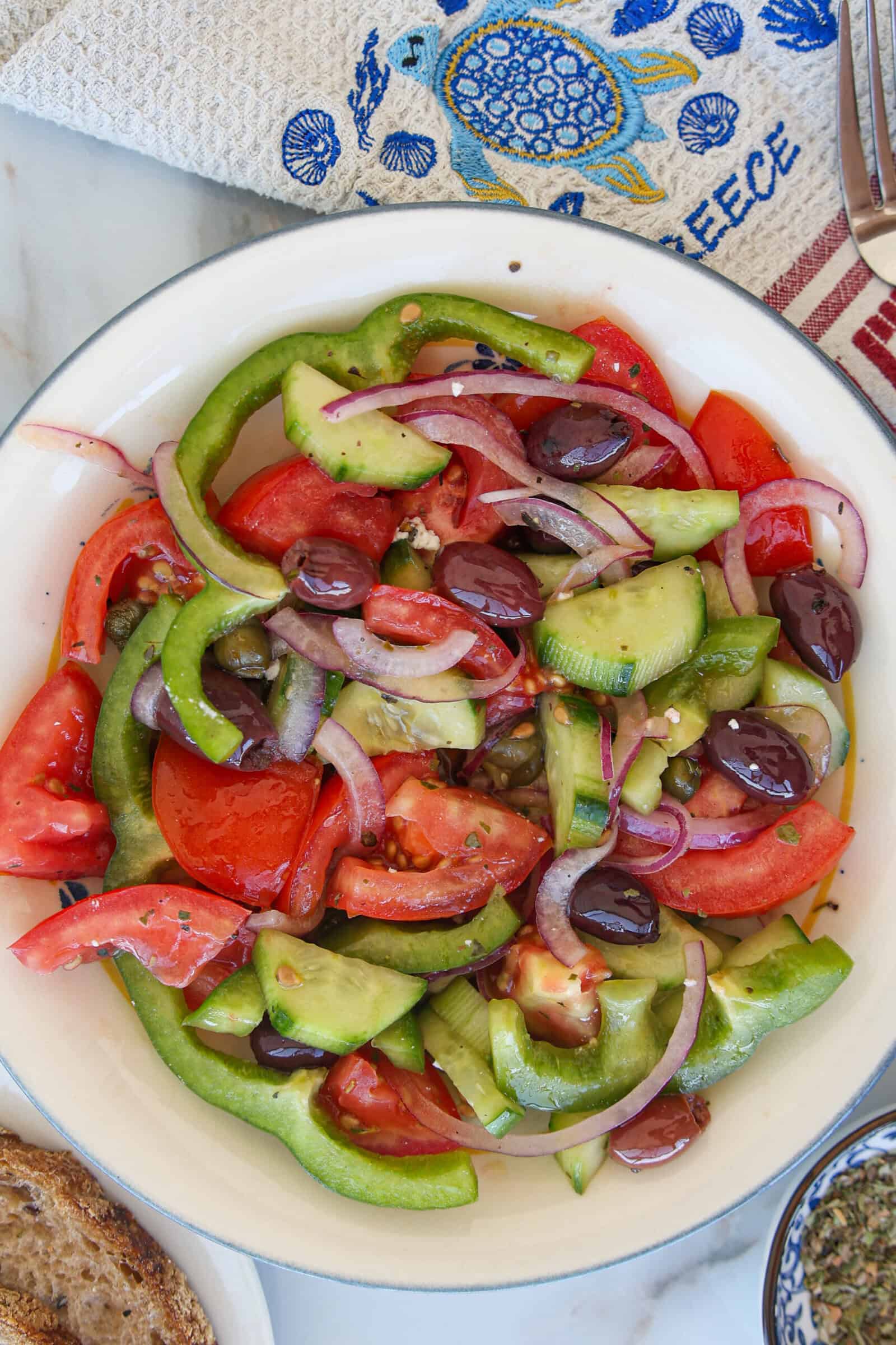 A bowl of Greek horiatiki with sliced tomatoes, cucumbers, green peppers, red onions, and Kalamata olives, on a white plate sitting on a marble surface, near a patterned napkin.