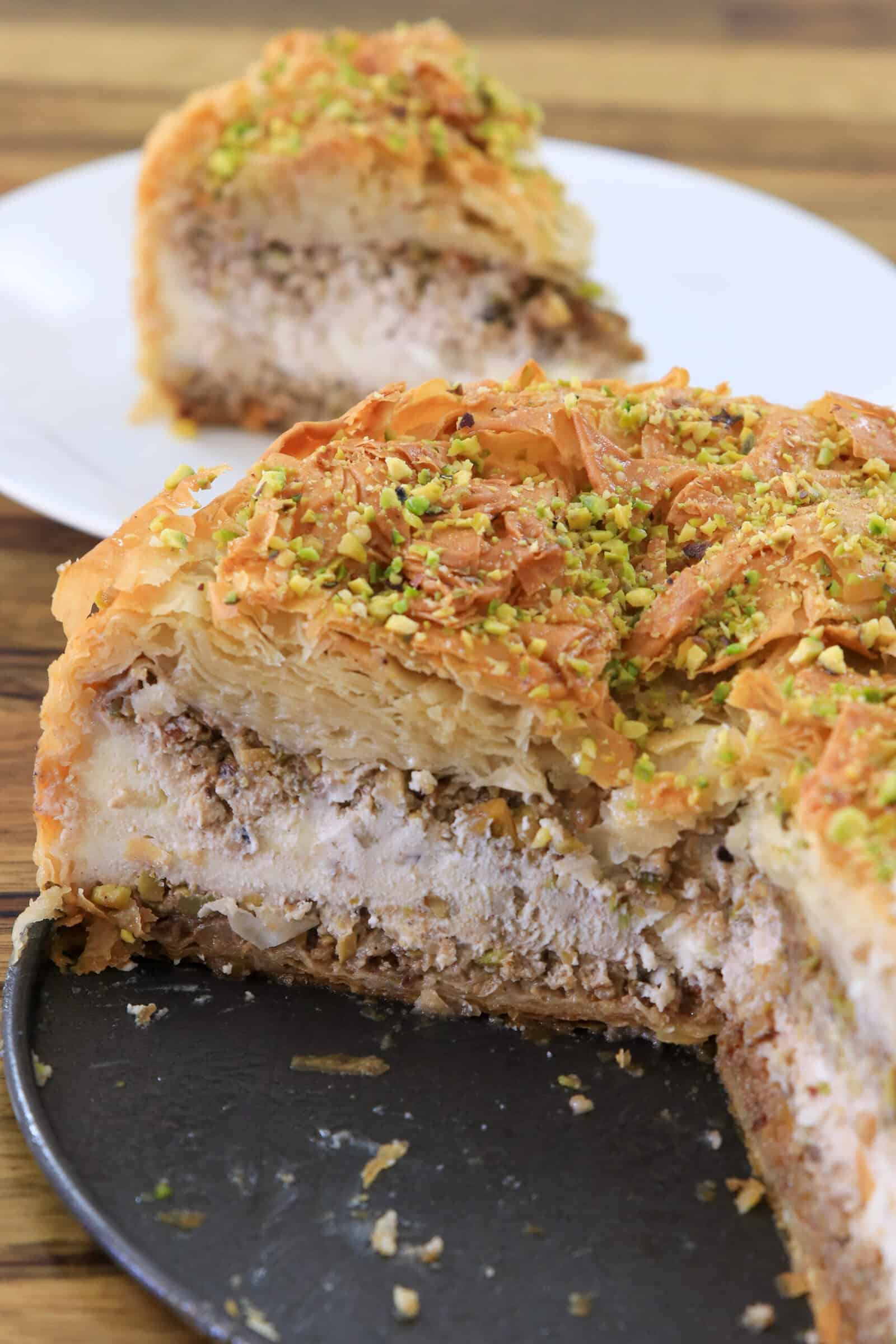 A close-up of a layered baklava cheesecake topped with chopped pistachios, with a slice served on a white plate in the background.