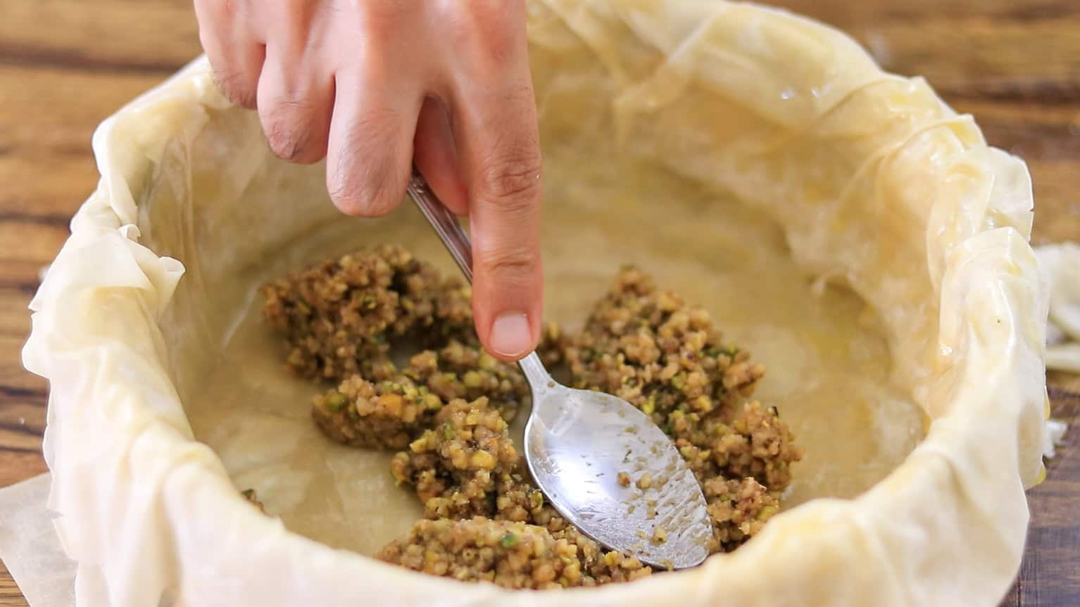 A hand uses a spoon to spread a ground meat and vegetable mixture onto a layer of filo dough in a round baking dish.
