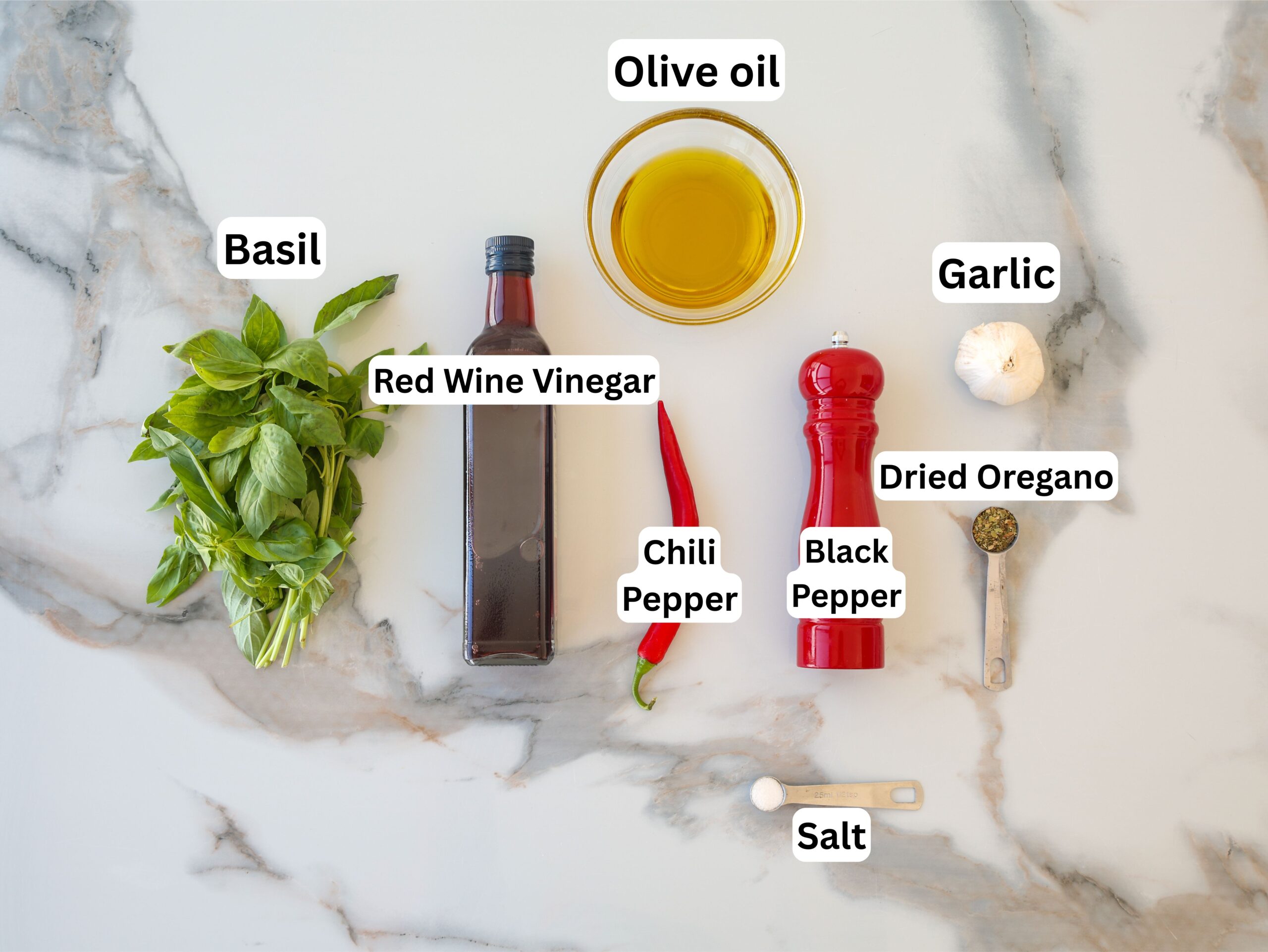 A marble surface with labeled ingredients: basil, red wine vinegar, olive oil, garlic, chili pepper, black pepper, dried oregano, and salt.