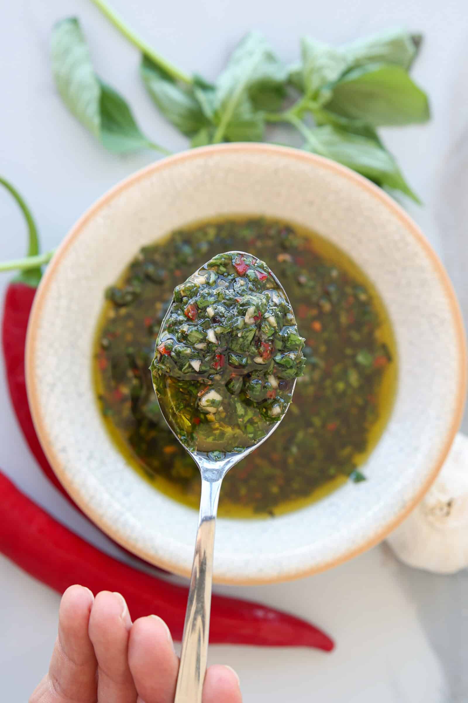 A spoon holds basil chimichurri sauce above a small bowl filled with the same sauce. Basil leaves, red chili peppers, and a garlic bulb rest nearby on a white surface.