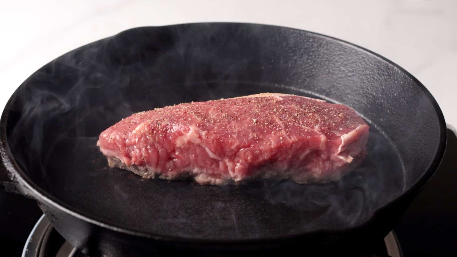 A thick, raw steak seasoned with pepper sizzles in a black cast iron skillet, with wisps of steam rising as it cooks on a stovetop.