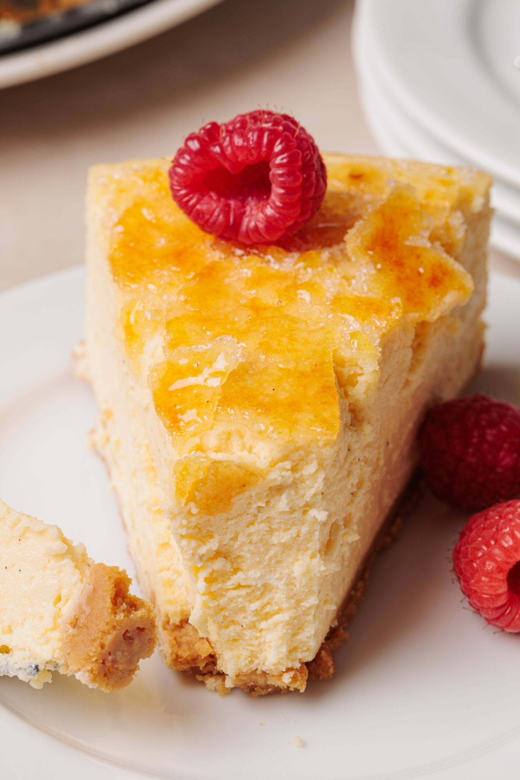 A slice of a creme brulee cheesecake topped with a single raspberry and a caramelized sugar crust sits on a white plate, with a couple of loose raspberries beside it.