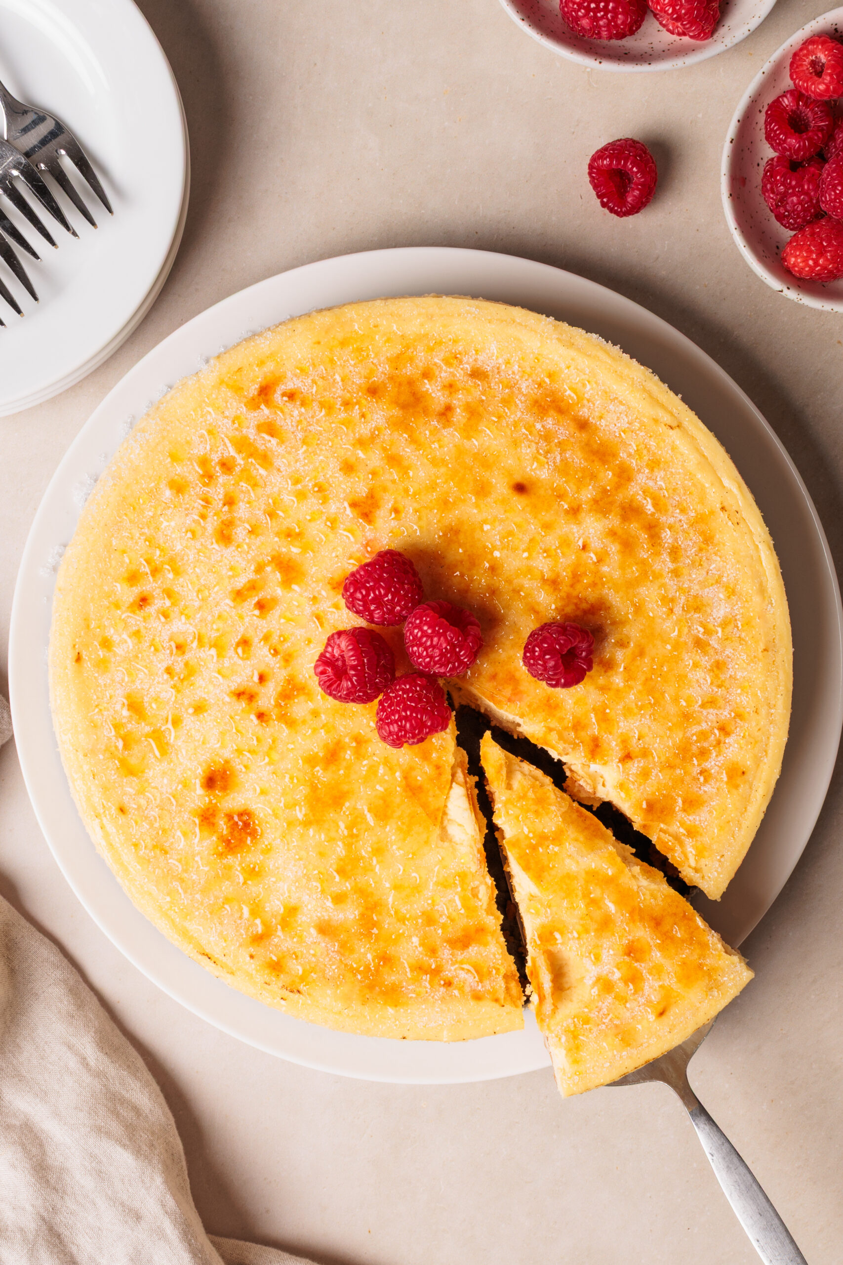 A creme brulee cheesecake with a golden top sits on a white plate. A slice is being lifted out, and fresh raspberries are placed in the center. Bowls of raspberries and a plate with forks are nearby.