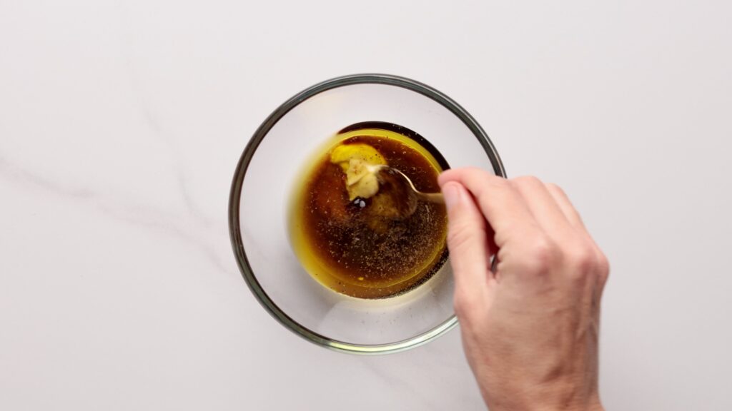 A hand stirs a mixture of oil, mustard, and seasonings in a clear glass bowl on a white surface.