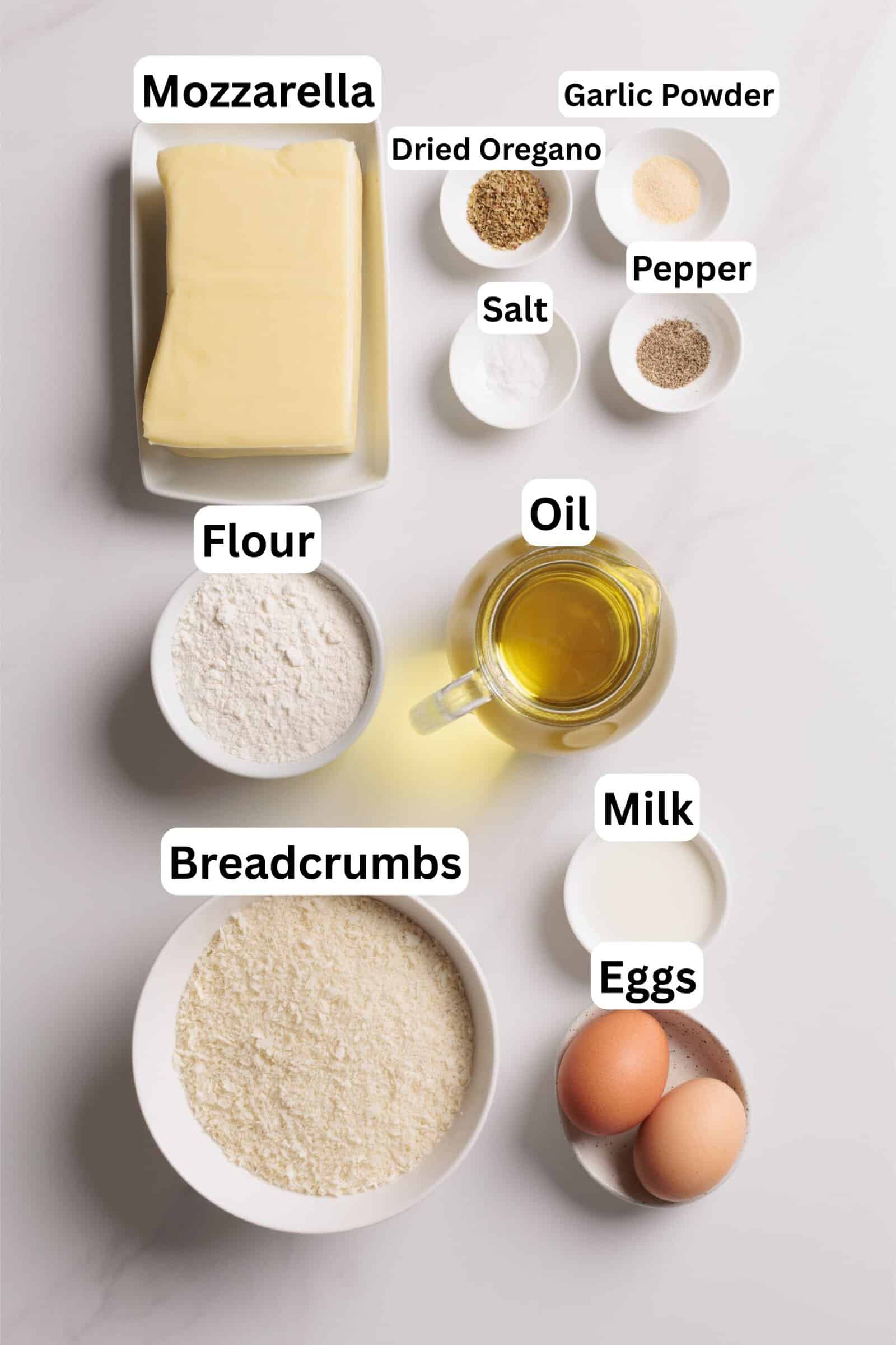 Ingredients for mozzarella sticks on a white surface: block of mozzarella, flour, breadcrumbs, two eggs, a cup of oil, bowls of dried oregano, garlic powder, salt, pepper, and a small bowl of milk.