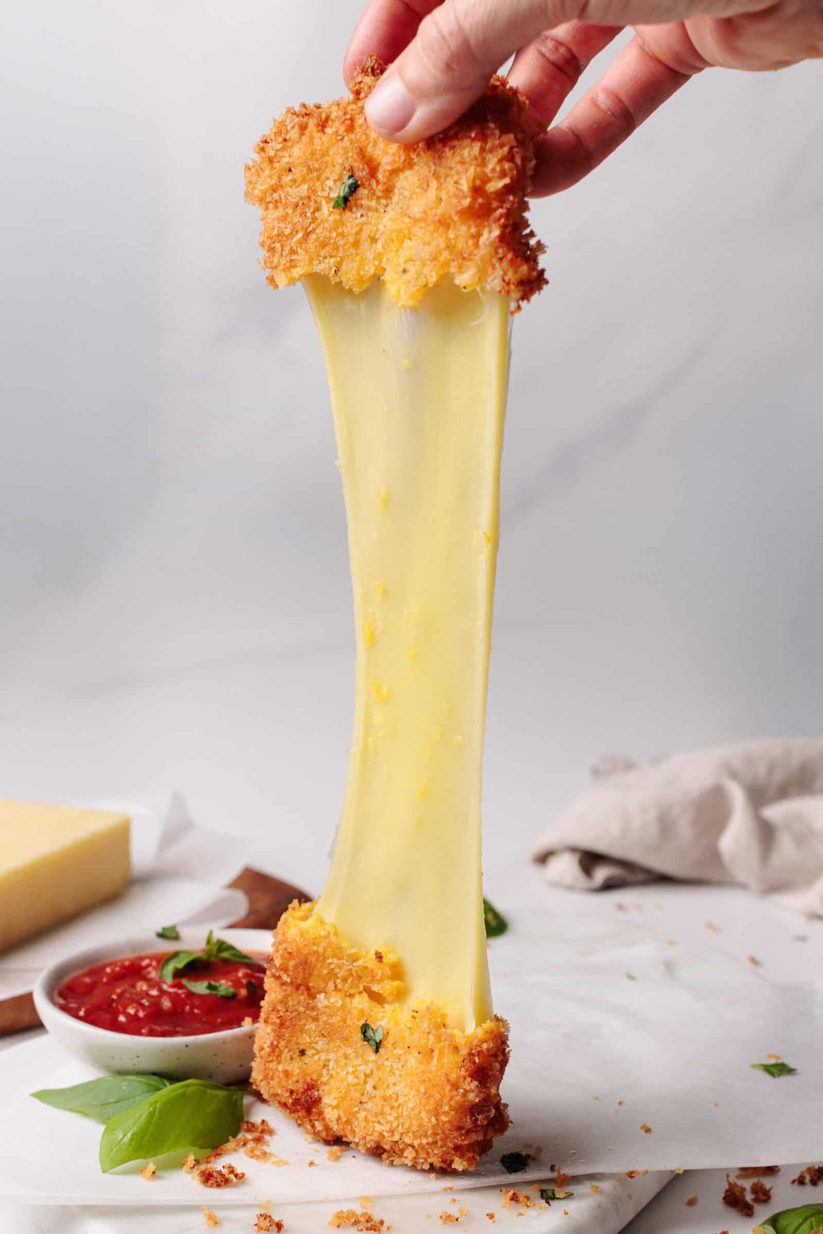 A hand pulls apart a breaded fried mozzarella stick, stretching melted cheese. Marinara sauce and basil leaves are on the table, along with a block of cheese in the background.