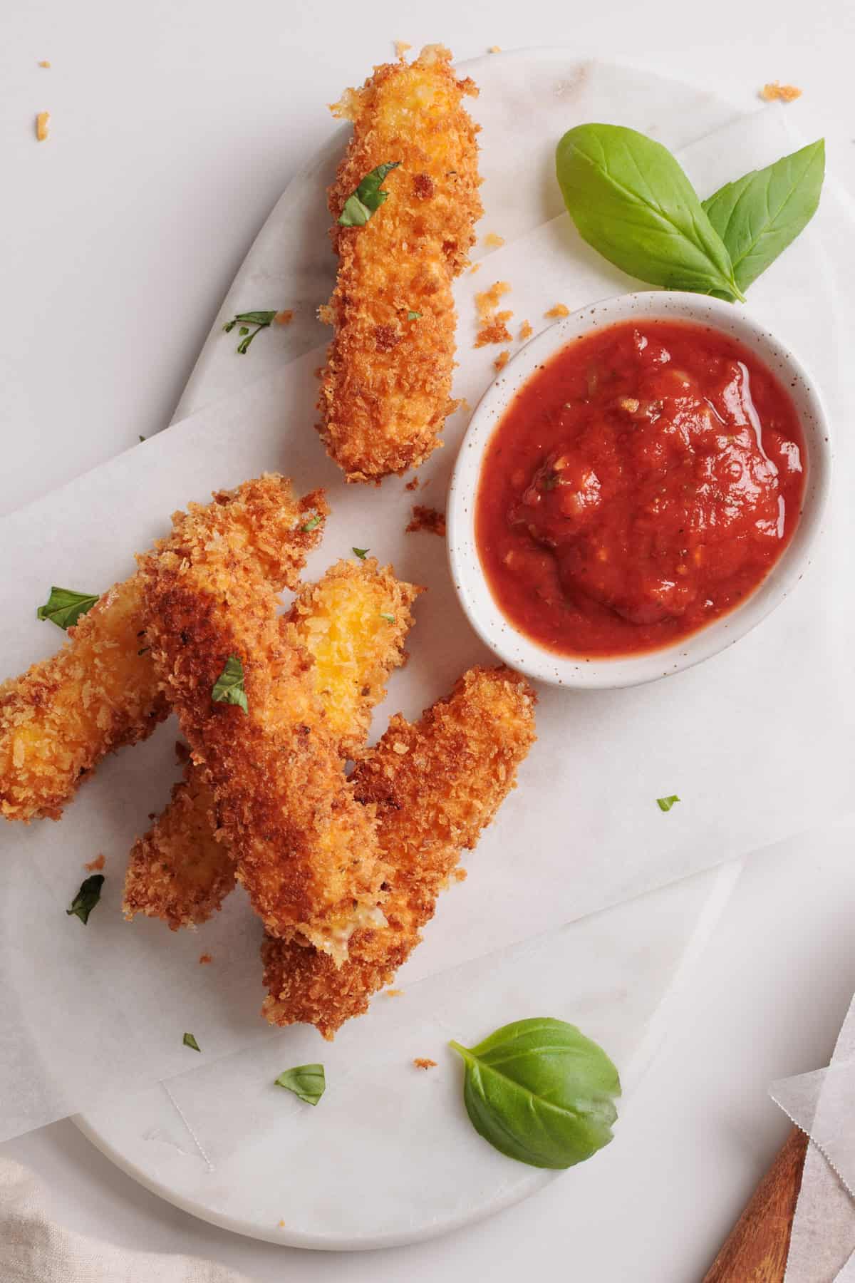 Golden breaded mozzarella sticks on a white platter with a small bowl of marinara sauce. Fresh basil leaves are scattered around the platter for garnish.