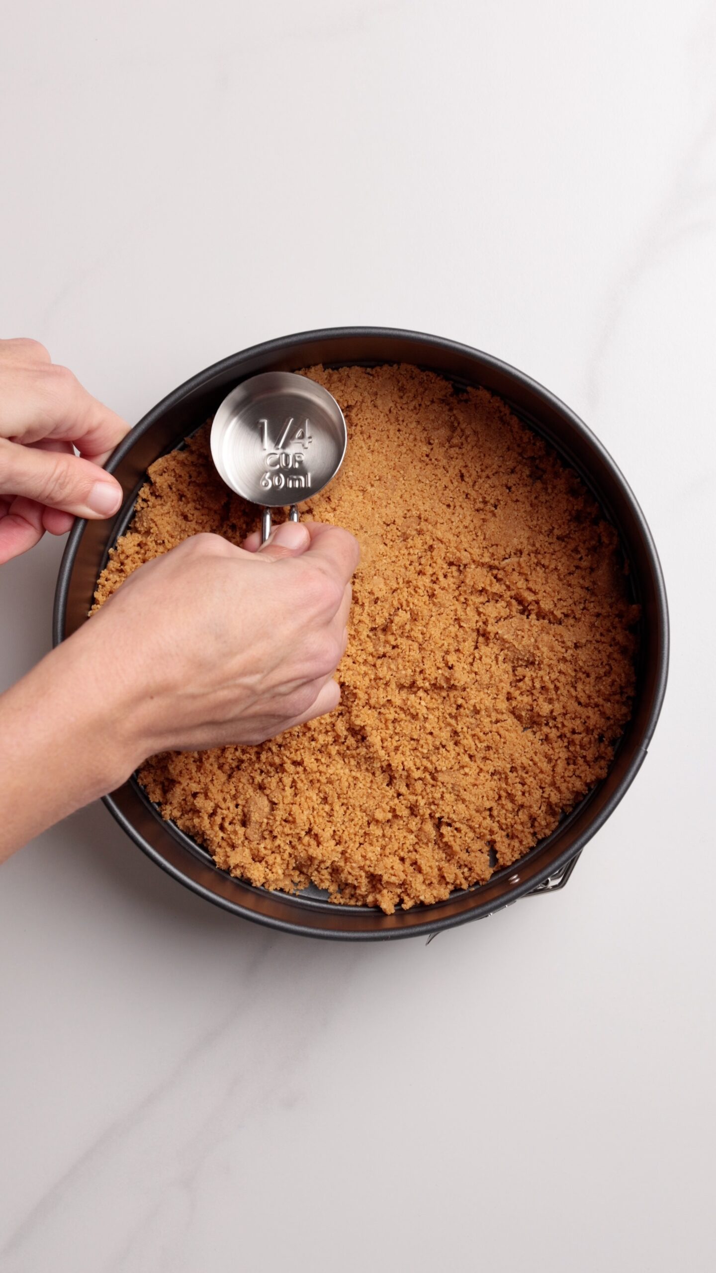 Hands pressing graham cracker crust into a round pan using a metal measuring cup on a light surface.