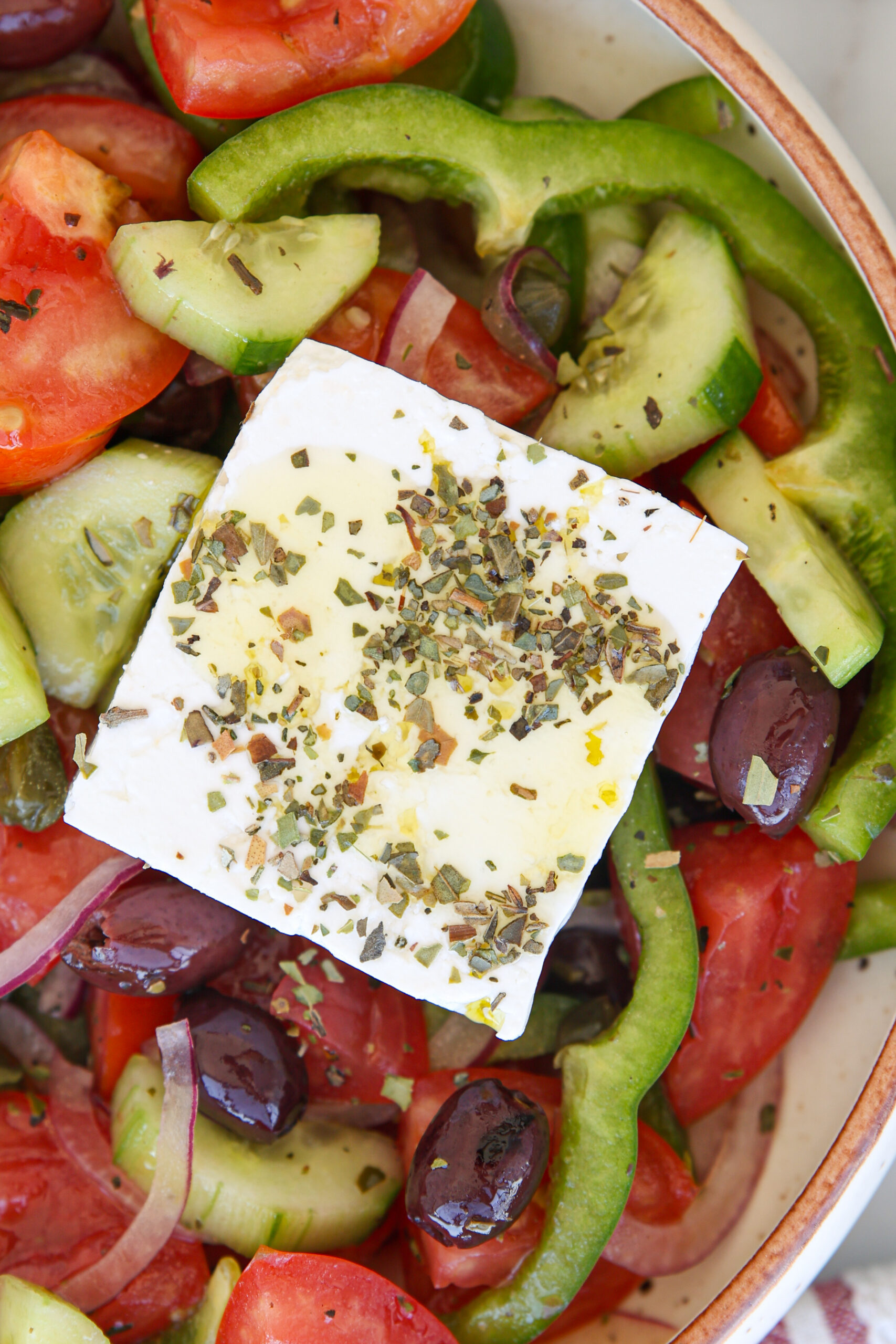 A bowl of Greek salad with sliced tomatoes, cucumbers, green peppers, red onions, olives, and a large square of feta cheese topped with herbs and olive oil.