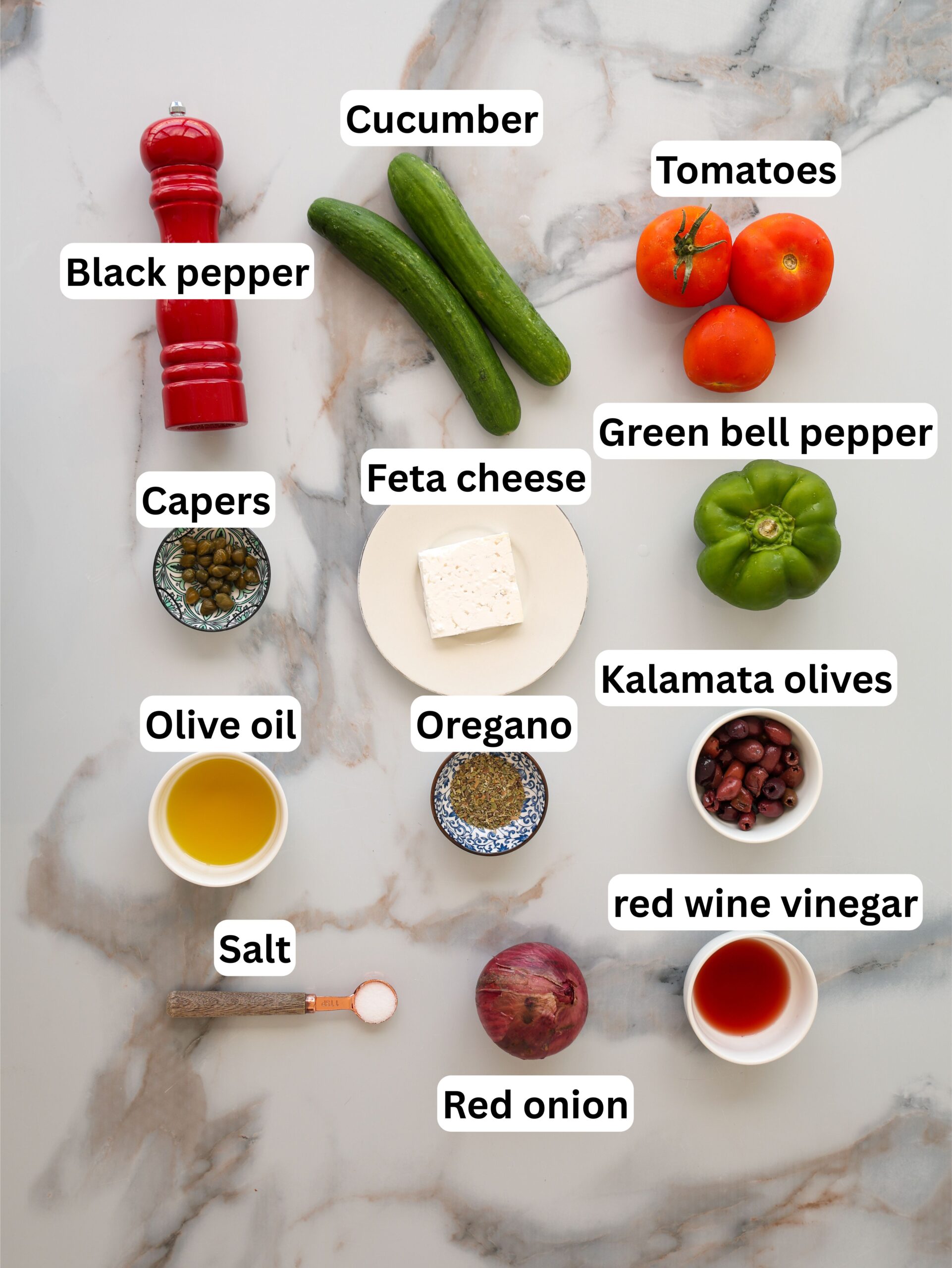 Ingredients for a Greek salad are arranged on a marble surface: cucumber, tomatoes, green bell pepper, feta cheese, Kalamata olives, capers, red onion, olive oil, red wine vinegar, black pepper, salt, and oregano.