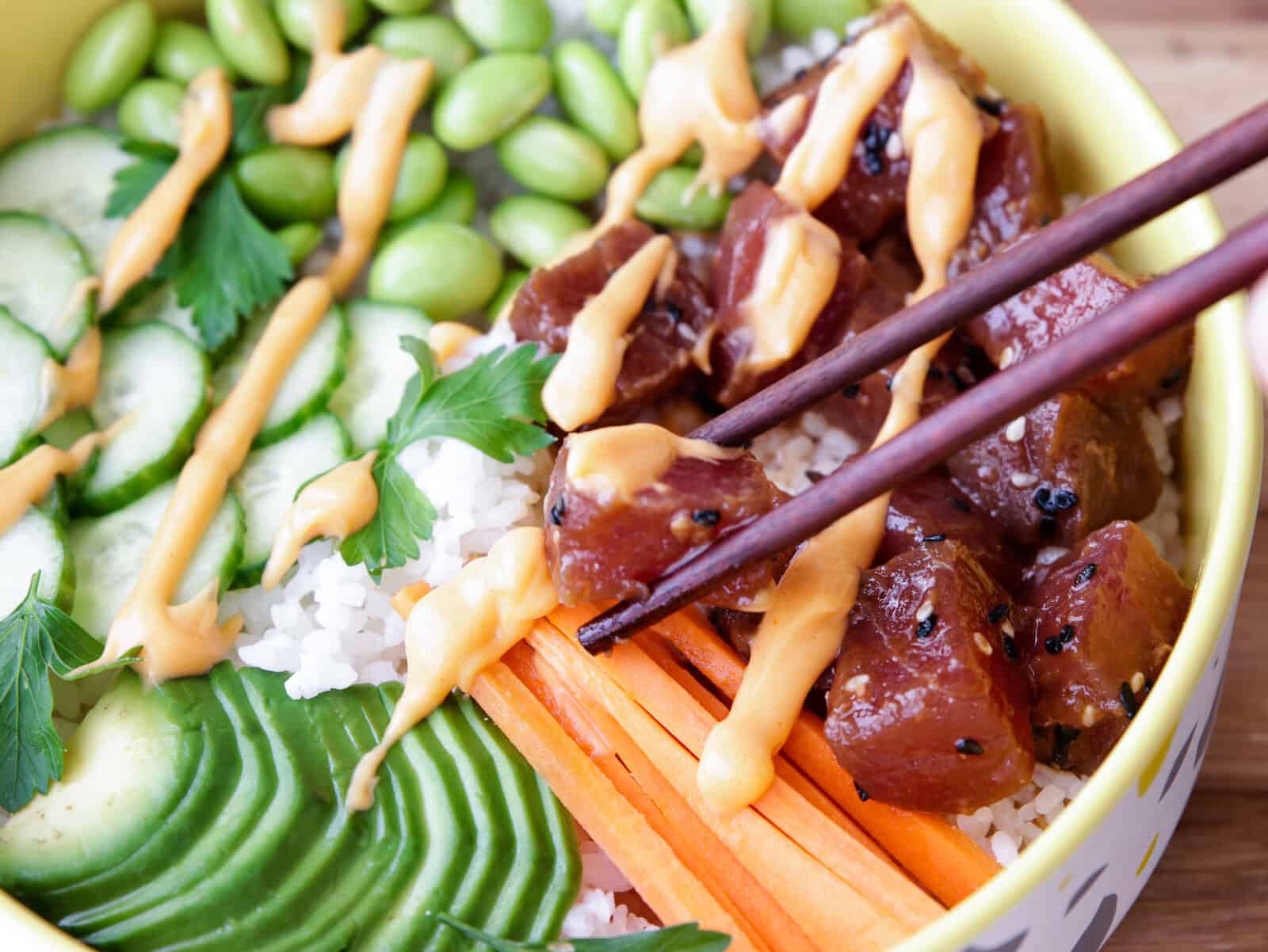 A close-up of a tuna poke bowl with rice, marinated fish, cucumber, edamame, carrot sticks, avocado, parsley, and a drizzle of orange sauce. Chopsticks are picking up a piece of fish.