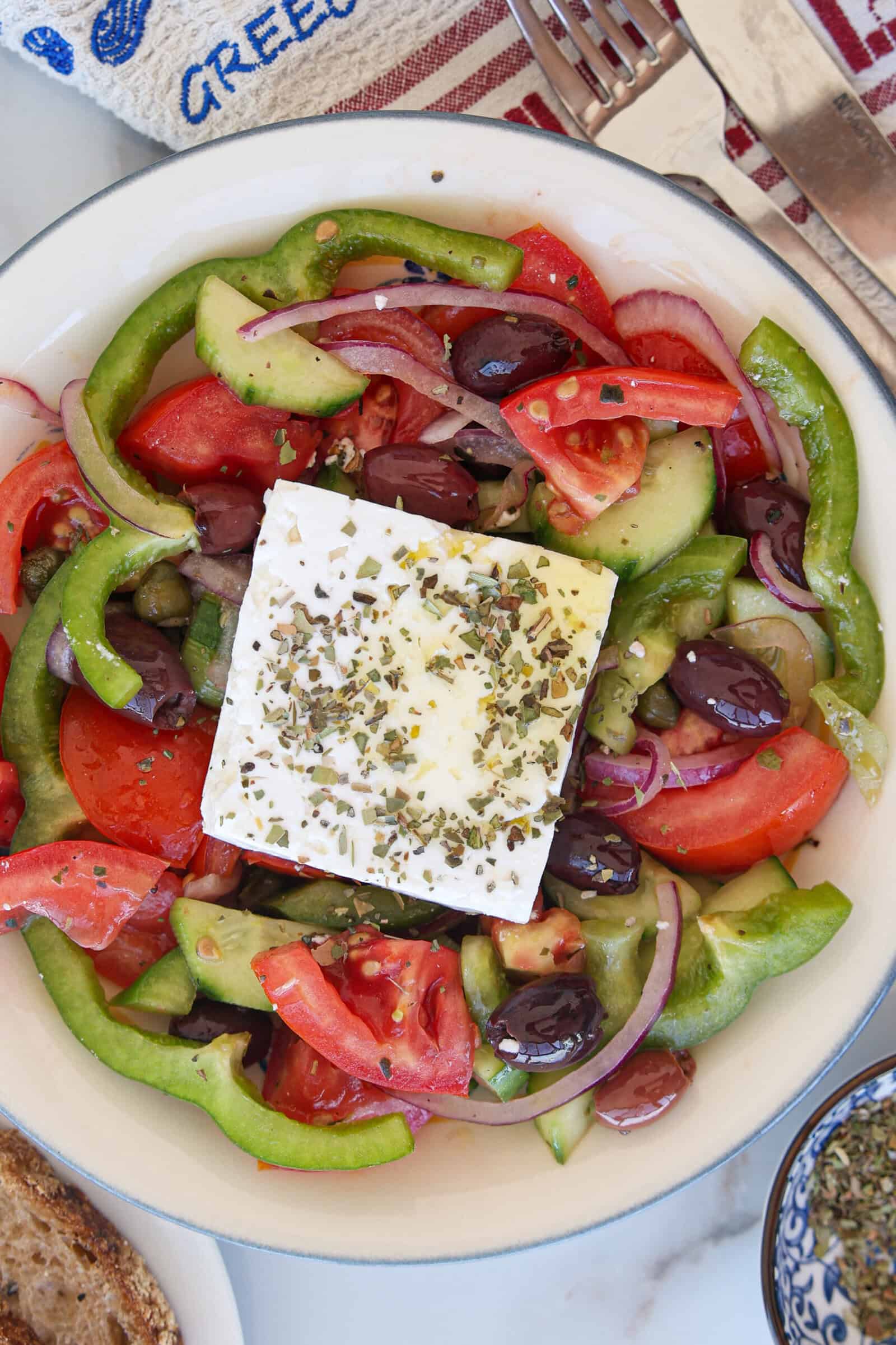 An authentic Greek salad with sliced tomatoes, cucumbers, green peppers, red onions, olives, and a large block of feta cheese topped with herbs, served in a white bowl.
