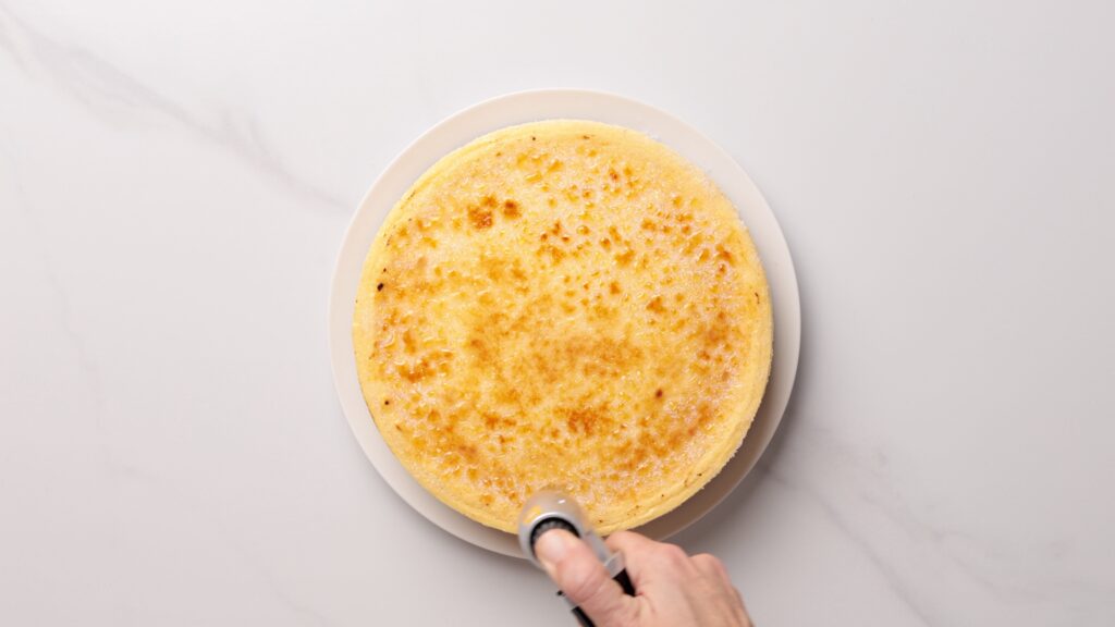 A hand holds a torch to brûlée the top of a round, yellow cake on a white plate, set on a light marble surface.