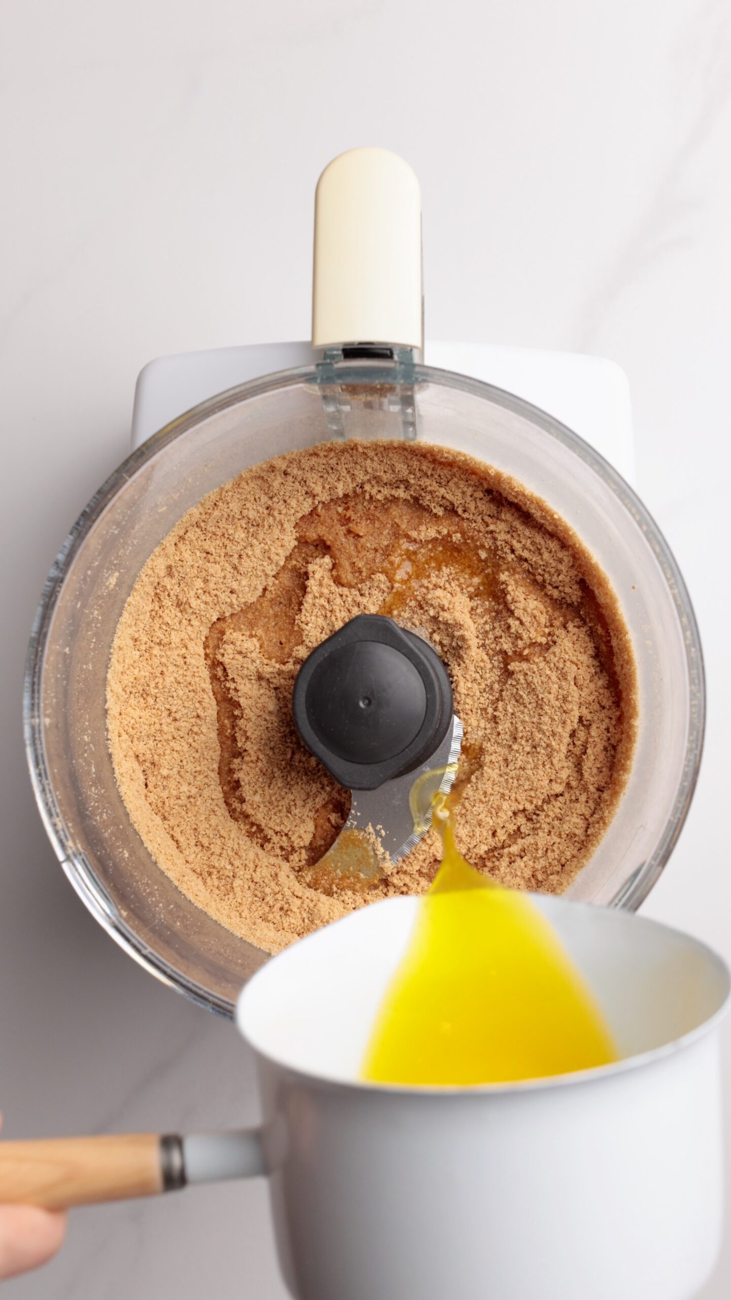 A food processor with crushed graham crackers inside, with melted butter being poured in from a white saucepan. The scene is set on a light-colored countertop.