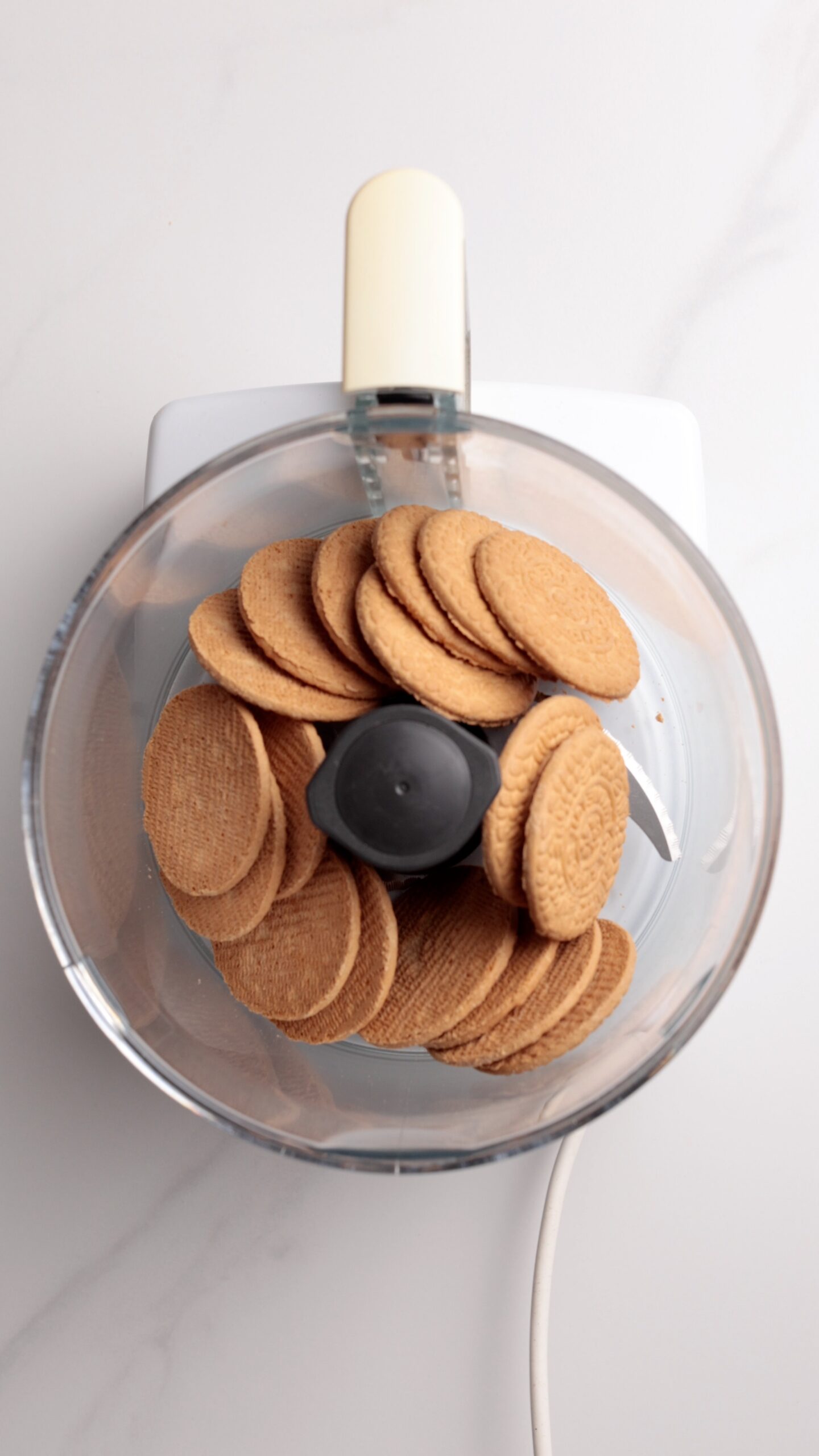 Overhead view of a food processor filled with whole round cookies, ready to be blended on a white countertop.
