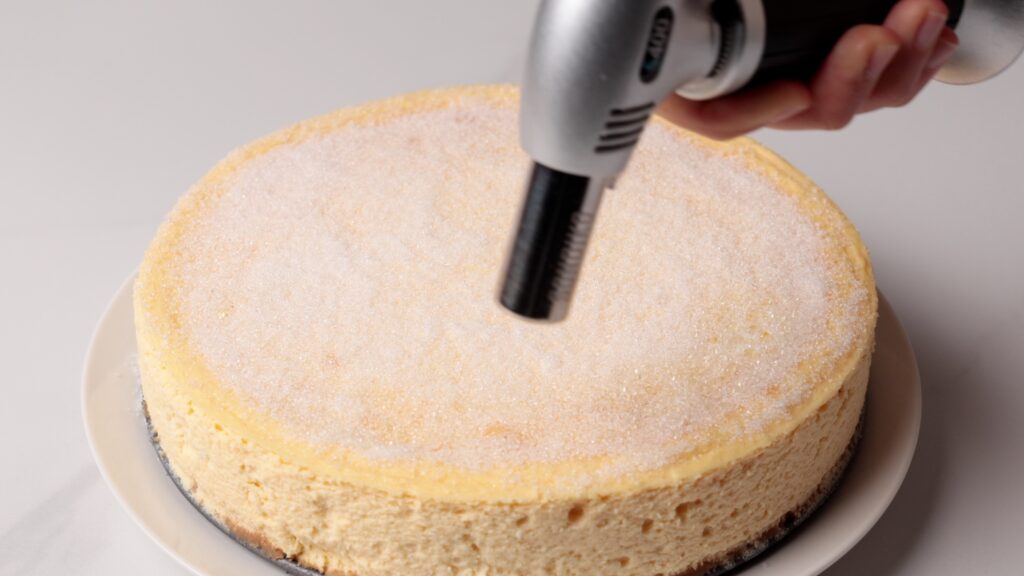 A hand uses a kitchen torch to caramelize a layer of sugar on top of a round cheesecake sitting on a white plate.