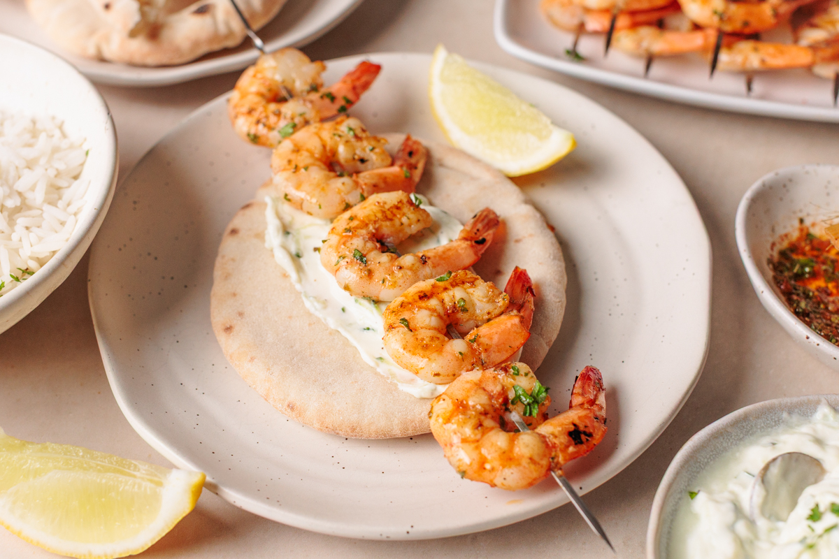 A skewer of grilled shrimp rests on pita bread with a layer of creamy sauce. Lemon wedges, rice, and more shrimp skewers are arranged around the plate on a light table.