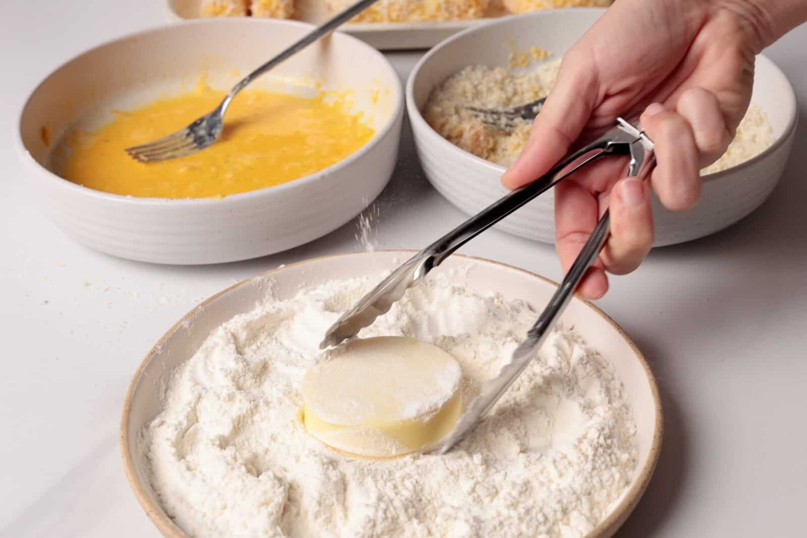 A hand uses tongs to coat a mozzarella slice in flour, with bowls of beaten egg and breadcrumbs nearby, preparing ingredients for cooking.