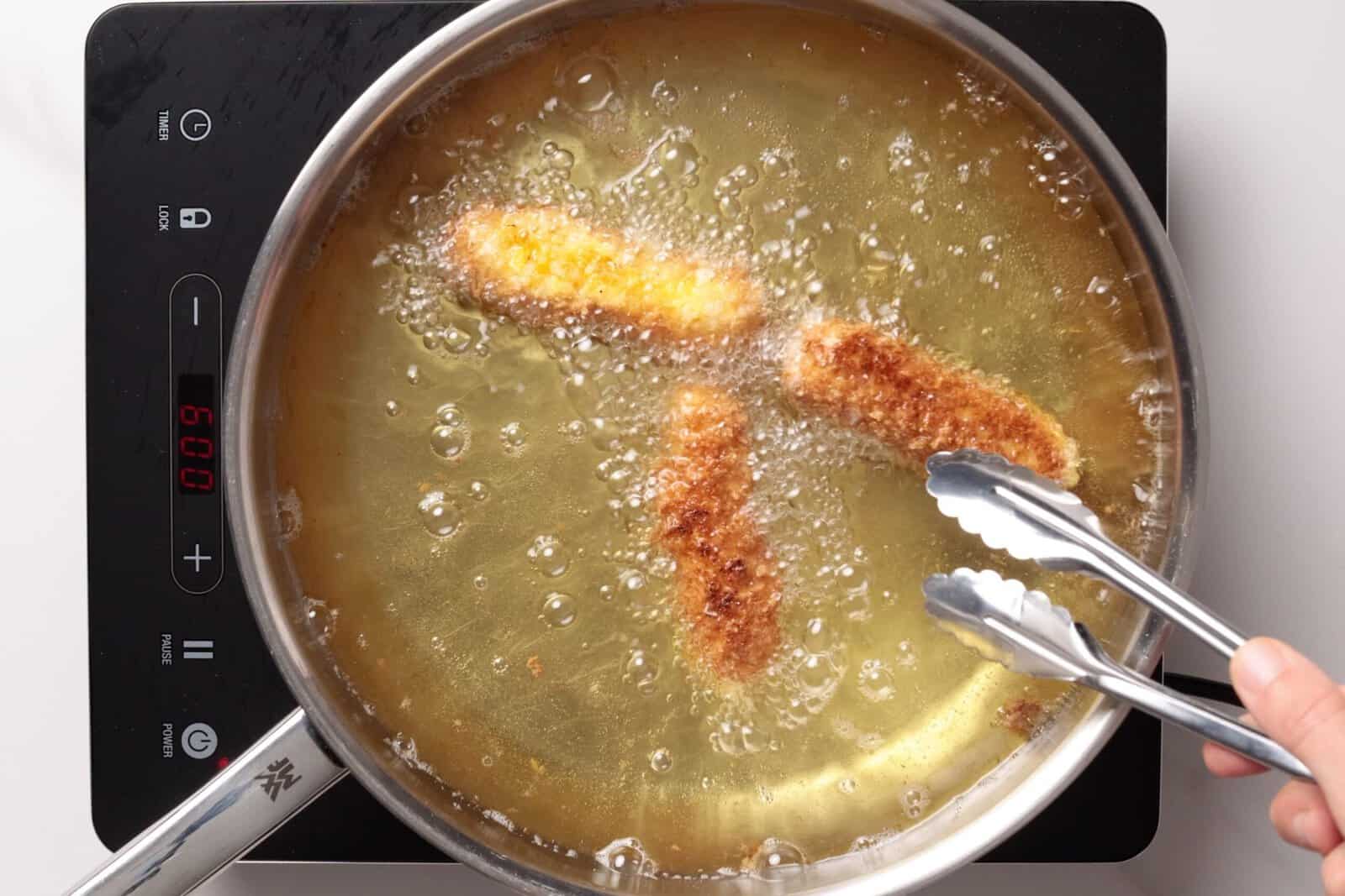 Breaded mozzarella sticks are being deep-fried in hot oil in a pan on an induction cooktop. A hand holds metal tongs, turning one of the pieces. Bubbles surround the frying food.