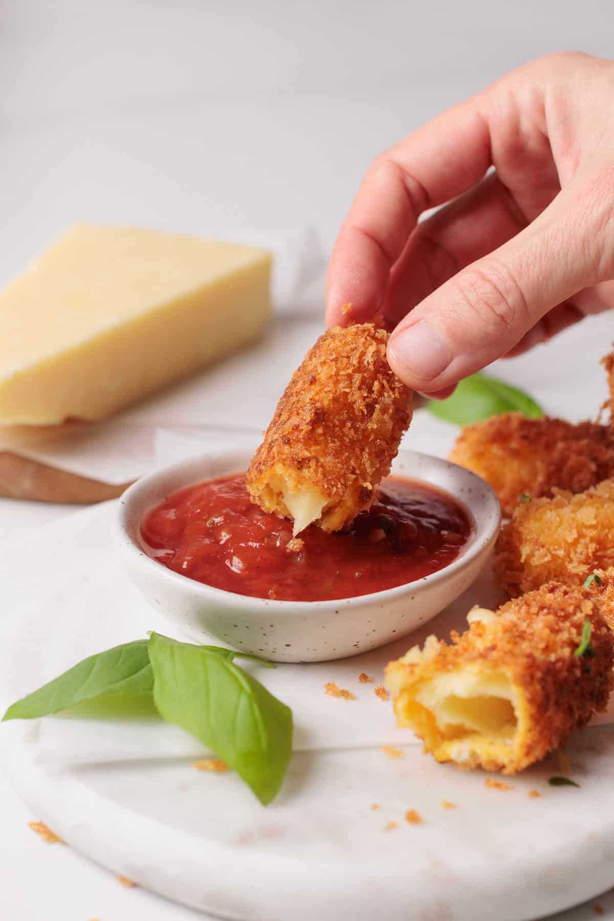 A hand dips a crispy fried mozzarella stick into a bowl of marinara sauce. Other mozzarella sticks, a wedge of cheese, and fresh basil leaves are nearby on a white marble surface.
