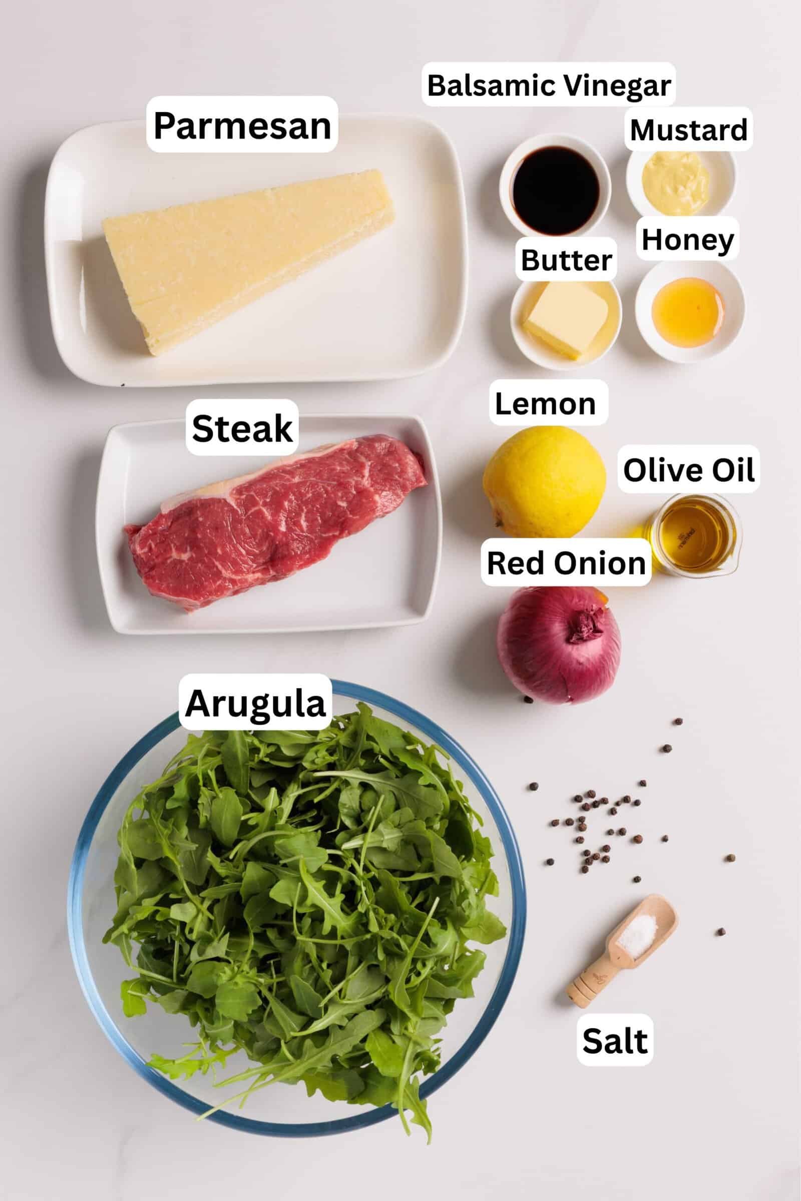 Flat lay of ingredients labeled: wedge of parmesan, balsamic vinegar, mustard, butter, honey, lemon, olive oil, steak, red onion, bowl of arugula, salt, and peppercorns on a white surface.