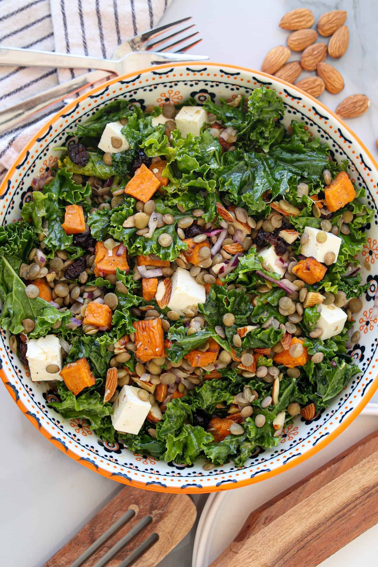 A colorful Kale Lentil Salad With Feta and Sweet Potatoes, with some almonds, red onion, and raisins in a patterned bowl, with utensils, almonds, and a striped napkin nearby.