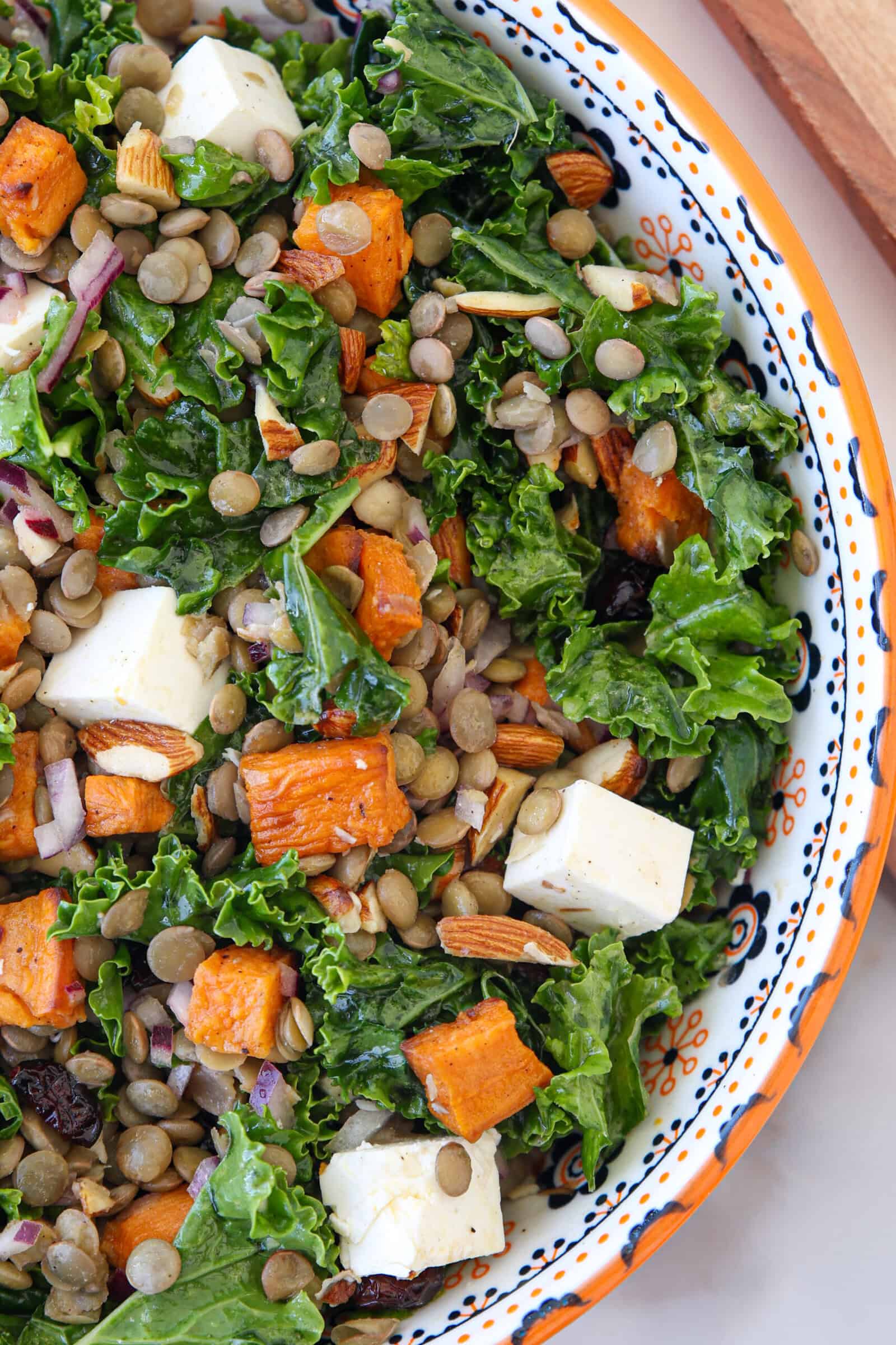 A colorful kale lentil salad with kale, roasted sweet potatoes, lentils, cubed feta cheese, chopped almonds, and red onions in a decorative bowl.
