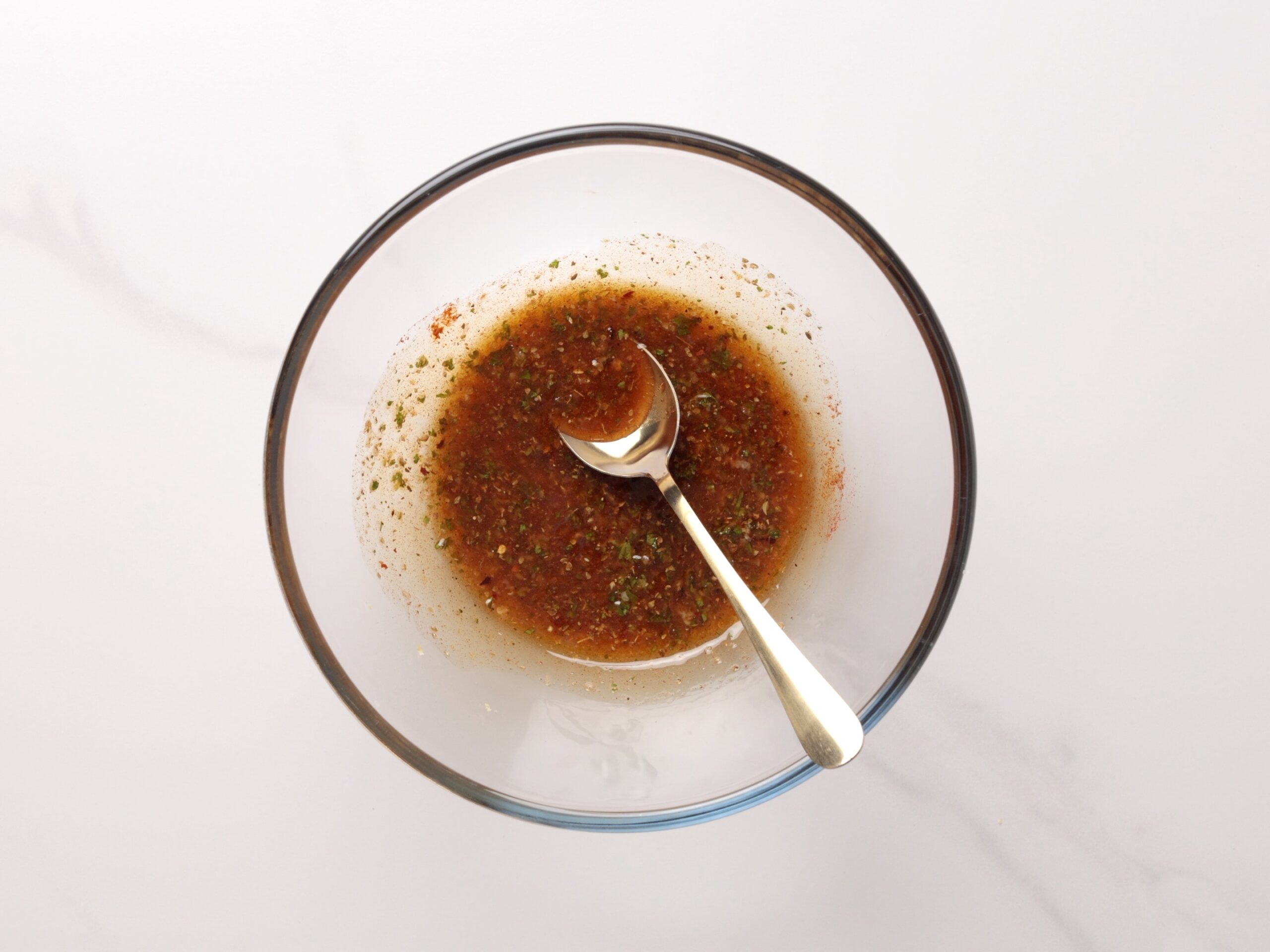 A clear glass bowl with a metal spoon, containing a marinade for shrimp skewers, sits on a white surface.