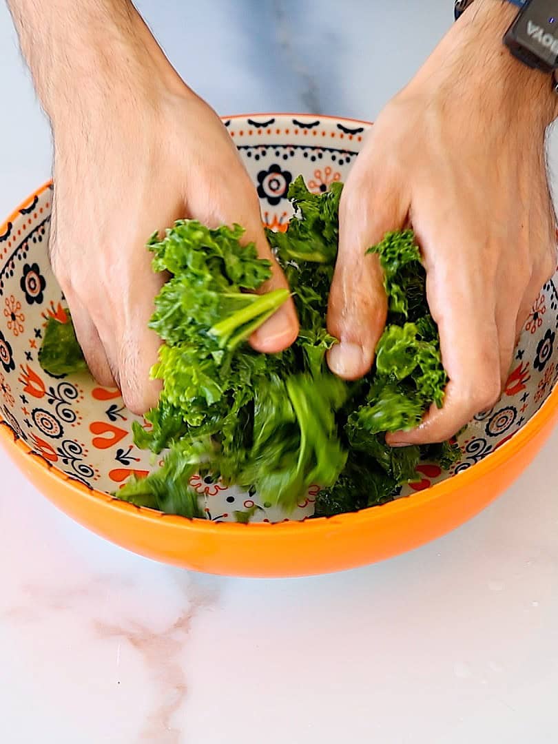Two hands massaging fresh kale leaves in a colorful orange bowl with decorative patterns, on a light surface.