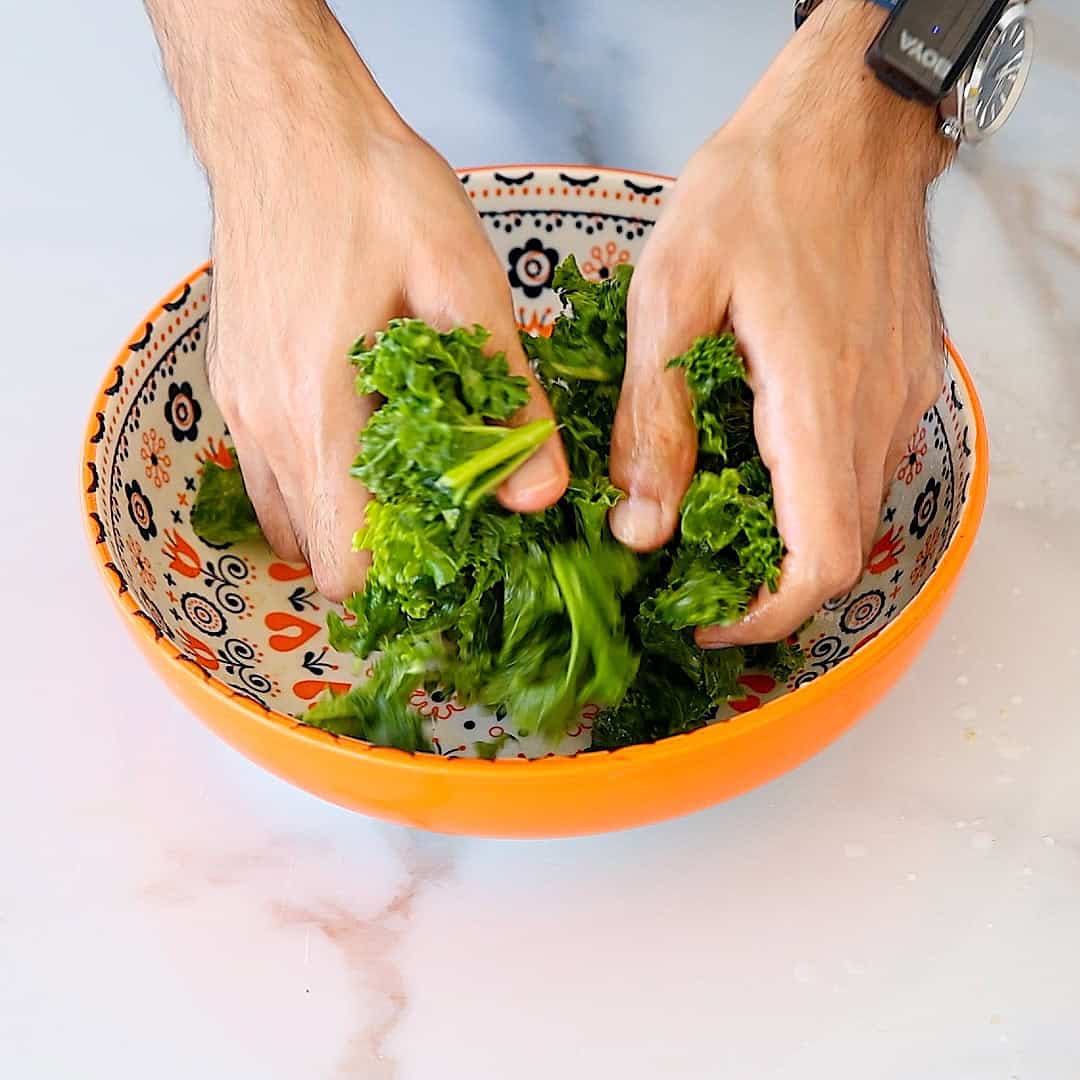Two hands massage leafy green kale in an orange patterned bowl on a white countertop.