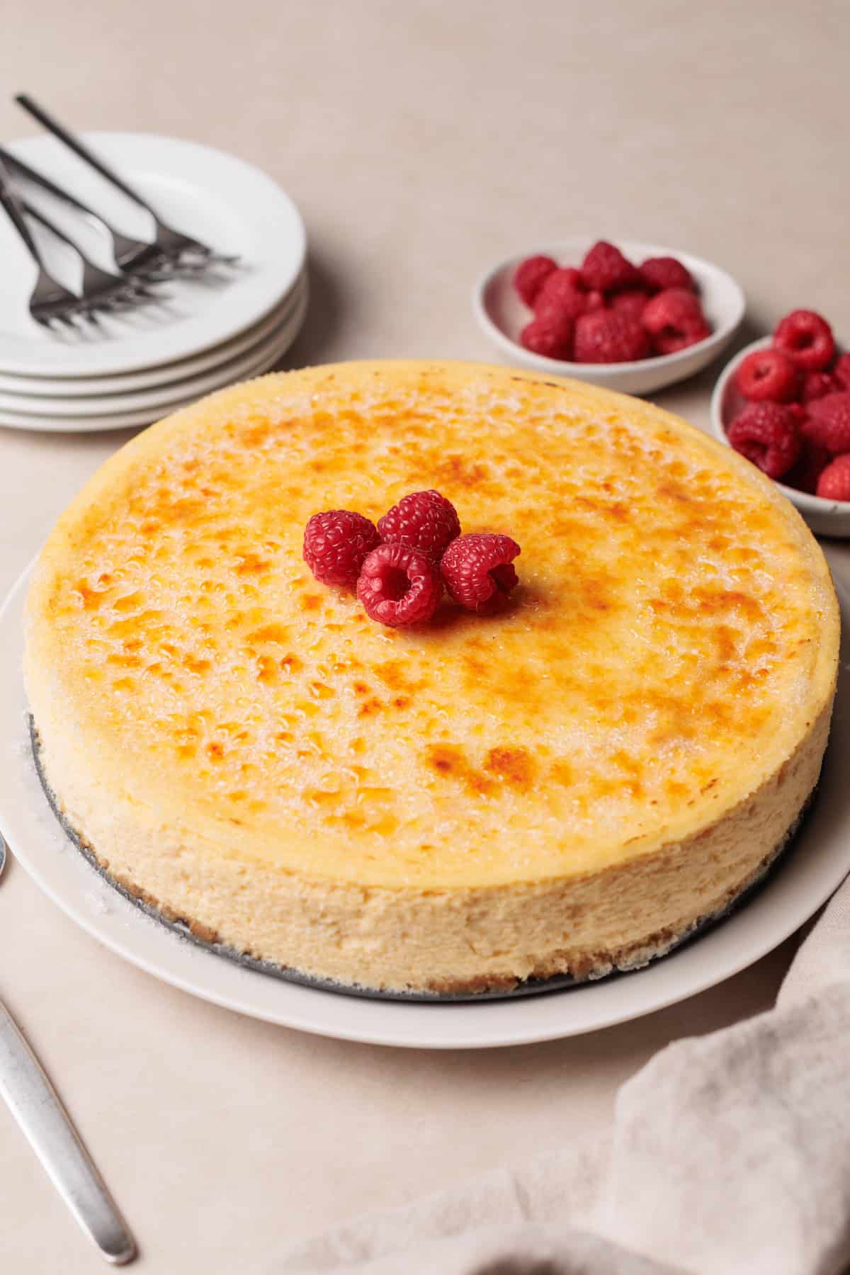 A cheesecake topped with fresh raspberries sits on a white plate. Nearby are bowls of raspberries, a stack of plates with forks, and a napkin. The scene is set on a light countertop.