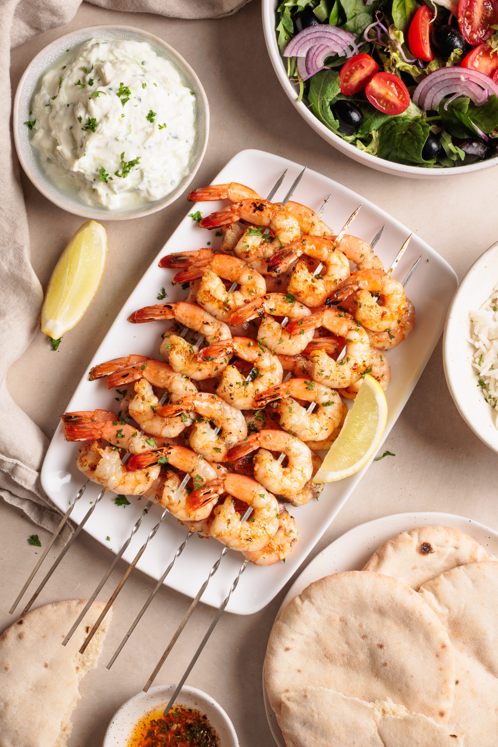 oven baked shrimp skewers on a white plate with lemon wedges, surrounded by pita bread, tzatziki sauce, a bowl of salad with olives and red onion, and a small dish of seasoned oil.