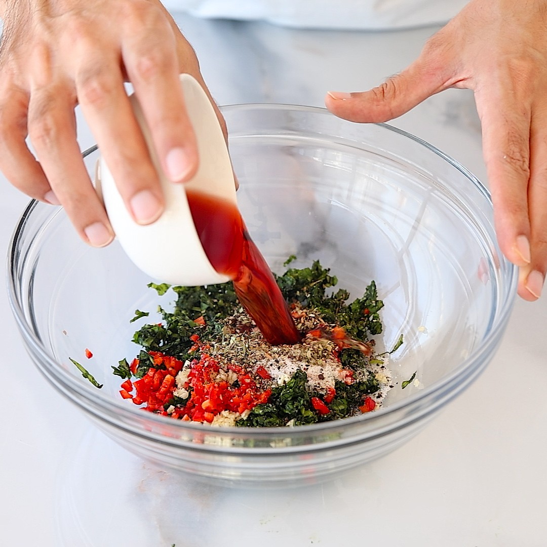 A person pours a brown liquid from a small bowl into a glass mixing bowl filled with chopped herbs, red peppers, and spices on a white surface.